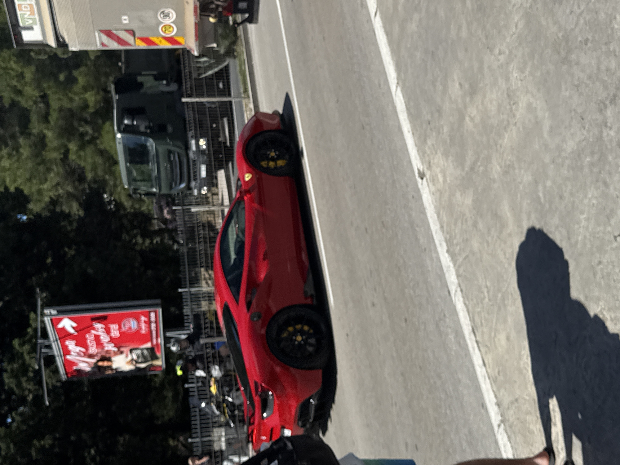 A red Ferrari is driving on a road in Budva, Montenegro. The car is in motion, viewed from the side. In the background, there is a green military truck and a large billboard advertising "Mega Stone". There are also trees and a metal fence. A person is partially visible in the foreground on the left, and another person is behind the fence on a motorcycle. The road surface is asphalt, with a white line marking the edge. The overall lighting suggests a sunny day.