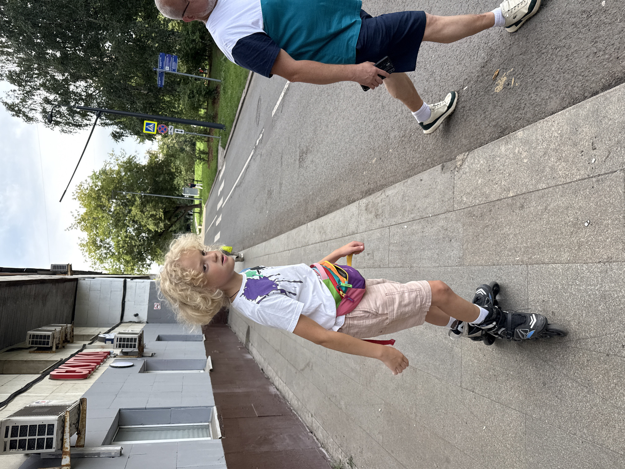 A young person with curly blonde hair is rollerblading on a sidewalk next to a street in Moscow, Russia. The person is wearing a white t-shirt with colorful paint splatters, light-colored shorts, and a brightly colored fanny pack around their waist. They are wearing black rollerblades. To the right of the sidewalk, an adult male wearing a white and teal shirt, dark shorts, white socks, and sneakers is walking on the asphalt. The building behind the rollerblader has gray and brown siding with several air conditioning units mounted on the exterior. Signs and trees are visible in the background.
