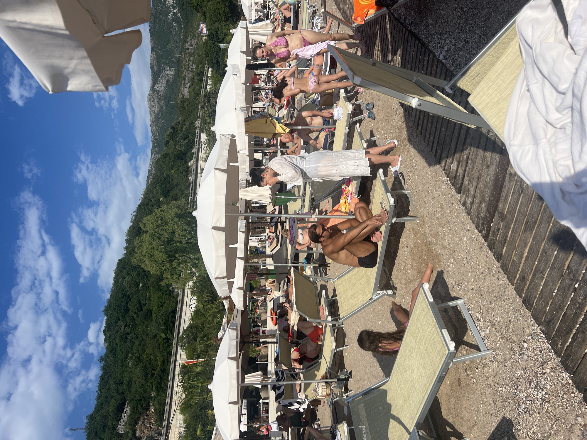 This media file captures a sun-drenched day at a beach in Kamenovo, Montenegro. The scene is populated with numerous people relaxing on lounge chairs, many positioned under white umbrellas. The beach appears to be a mix of sand and small pebbles.
The subjects are predominantly people in swimwear, engaged in typical beach activities like sunbathing and resting. Some are seen reading or simply enjoying the atmosphere. Lounge chairs and large white umbrellas dominate the foreground and middle ground, providing shade. In the background, a lush green hillside rises, dotted with trees, and a clear blue sky with scattered white clouds stretches above. The wooden boardwalk runs along the edge of the beach.
The time of day is clearly daytime, indicated by the bright sunlight casting distinct shadows, suggesting it's likely midday or early afternoon. The weather appears to be warm and sunny, ideal for a beach outing. The prominent hillside and the style of the beach setup are characteristic of a Mediterranean coastal location, consistent with Kamenovo, Montenegro. There is no visible text in the image. The overall mood conveyed is one of relaxation and leisure.