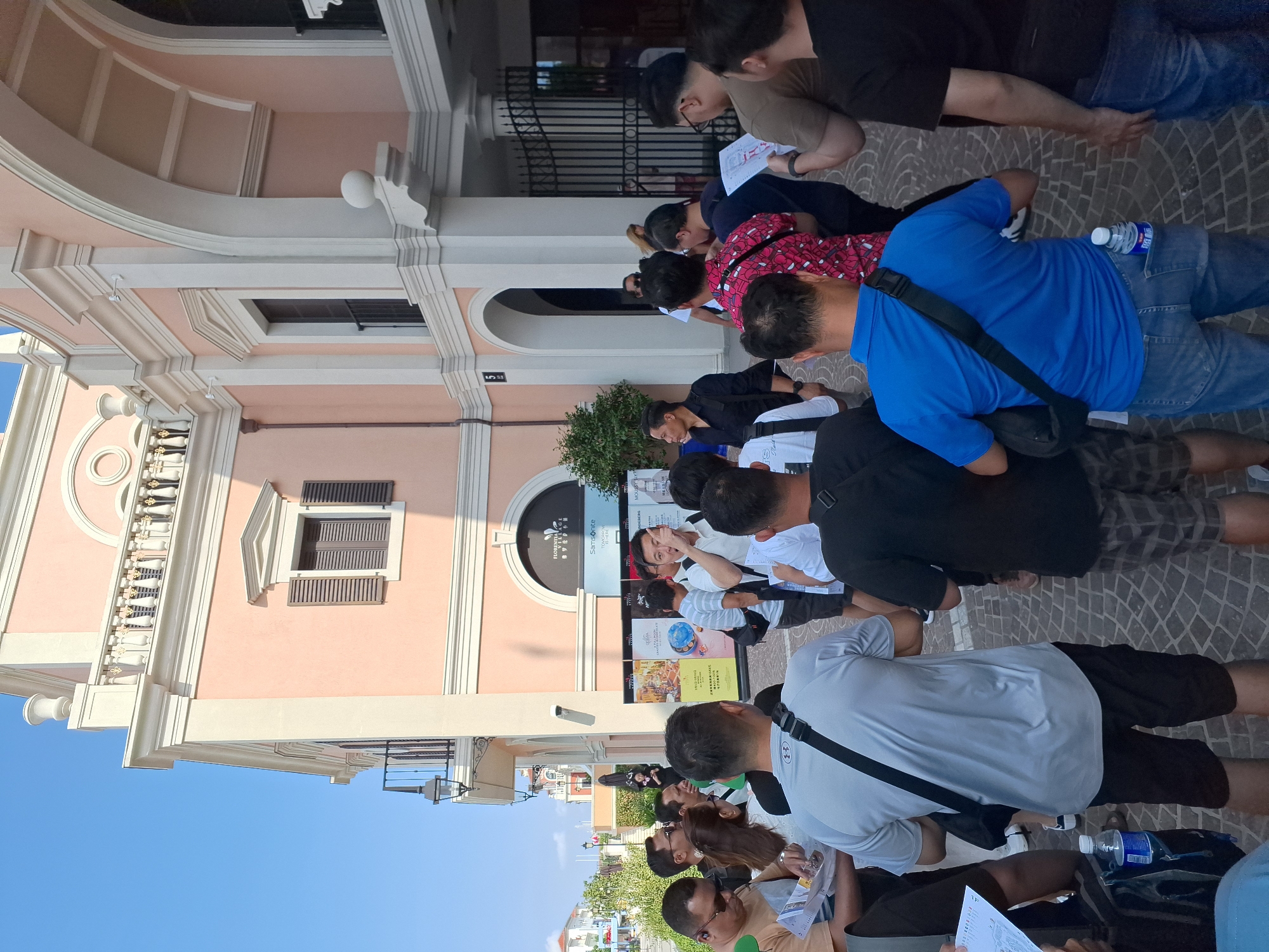 The image depicts a group of people gathered outdoors in front of a building with a light pink facade and white architectural trim. The building features arched doorways, balconies with decorative railings, and shuttered windows. Signs are visible on the building's exterior, including one with the text "FLORINTIAGE VILLAGE" and "SAMSONITE." The ground is paved with cobblestones. Several individuals, predominantly men, are standing and looking in various directions. Some are holding papers or maps. One man in a blue shirt appears to be gesturing while speaking. The scene suggests a public gathering or event. The environment is bright and sunny, indicating daytime.