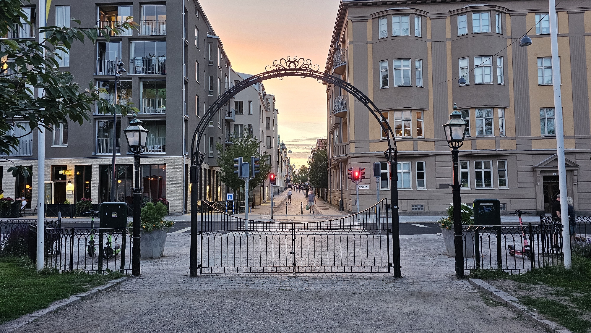 The image shows a street view in Linköping, Sweden, looking down a street lined with apartment buildings, framed by a decorative arched gateway and closed metal gates. The sky above is a warm gradient of orange and yellow, indicating either sunrise or sunset.
In the foreground, a paved pathway leads towards the closed gates. The gates are ornate, made of black wrought iron, with a decorative arch above them. To the left of the gates, a black lamppost stands beside a green bin and a small patch of grass. Further to the left, a building with a dark facade and multiple windows is visible, with a tree in the foreground casting shadows. To the right of the gates, another lamppost is visible, as is a large planter with greenery. A pink electric scooter is parked behind the fence to the right.
The street beyond the gates is paved with cobblestones and has a pedestrian crossing marked. Traffic lights are visible and illuminated red, indicating that traffic is stopped. Several people are walking on the sidewalk and crossing the street. The buildings on either side of the street are multi-story apartment blocks with many windows. The building on the right has a distinctive facade with vertical yellow and gray stripes.
The overall impression is one of urban tranquility at dusk or dawn, with the street appearing calm and orderly. The architectural style of the buildings suggests a European city setting.