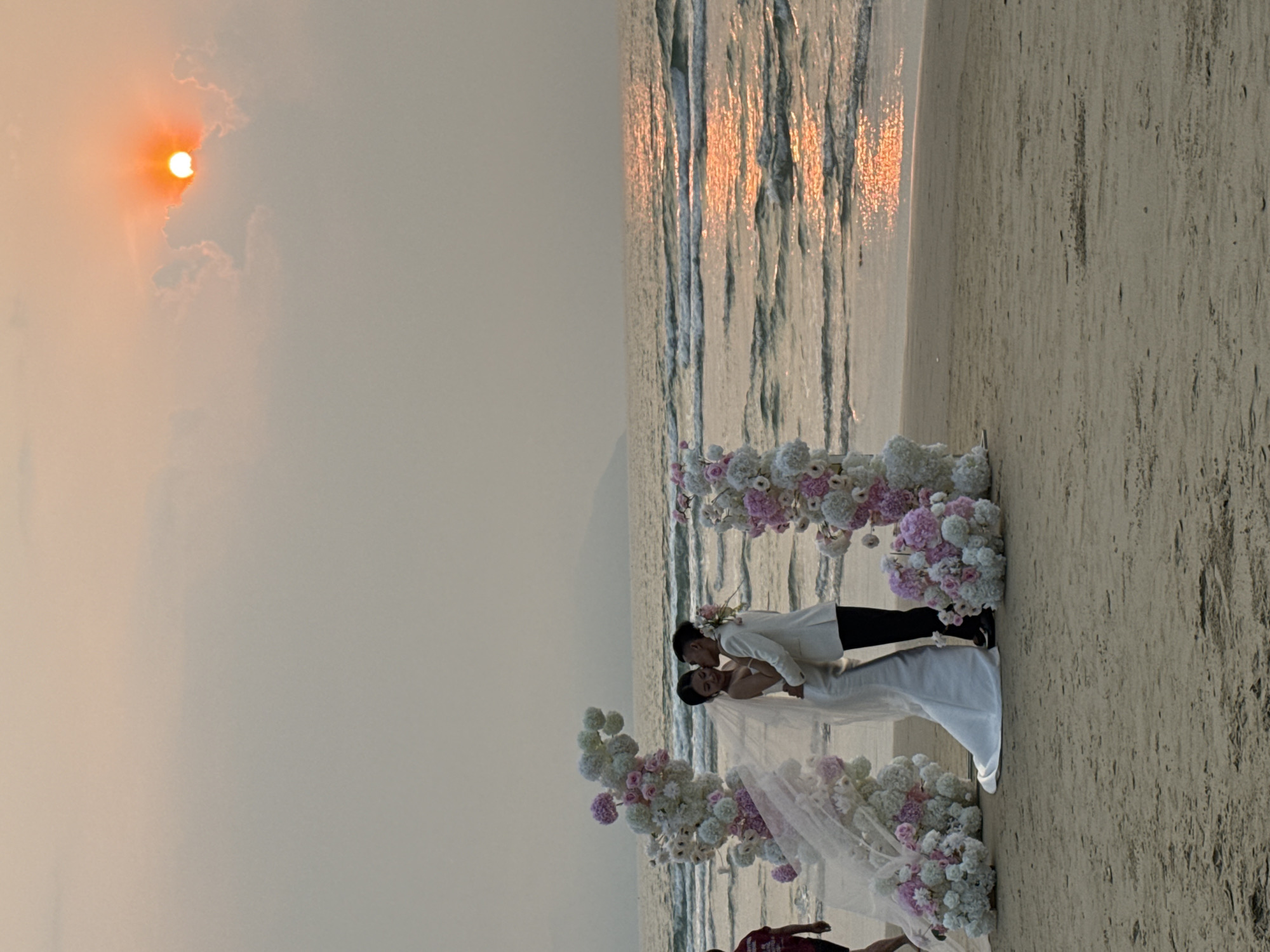 The image shows a wedding ceremony on a beach at sunset in Đà Nẵng, Vietnam. A couple is embracing in front of a floral arch on the sand near the water's edge. The groom is wearing a white suit and the bride is wearing a white wedding gown with a veil. The sun is setting in the background, casting a warm glow on the scene. The ocean waves are visible in the middle of the frame. A person wearing a red shirt is visible in the bottom center of the frame.
