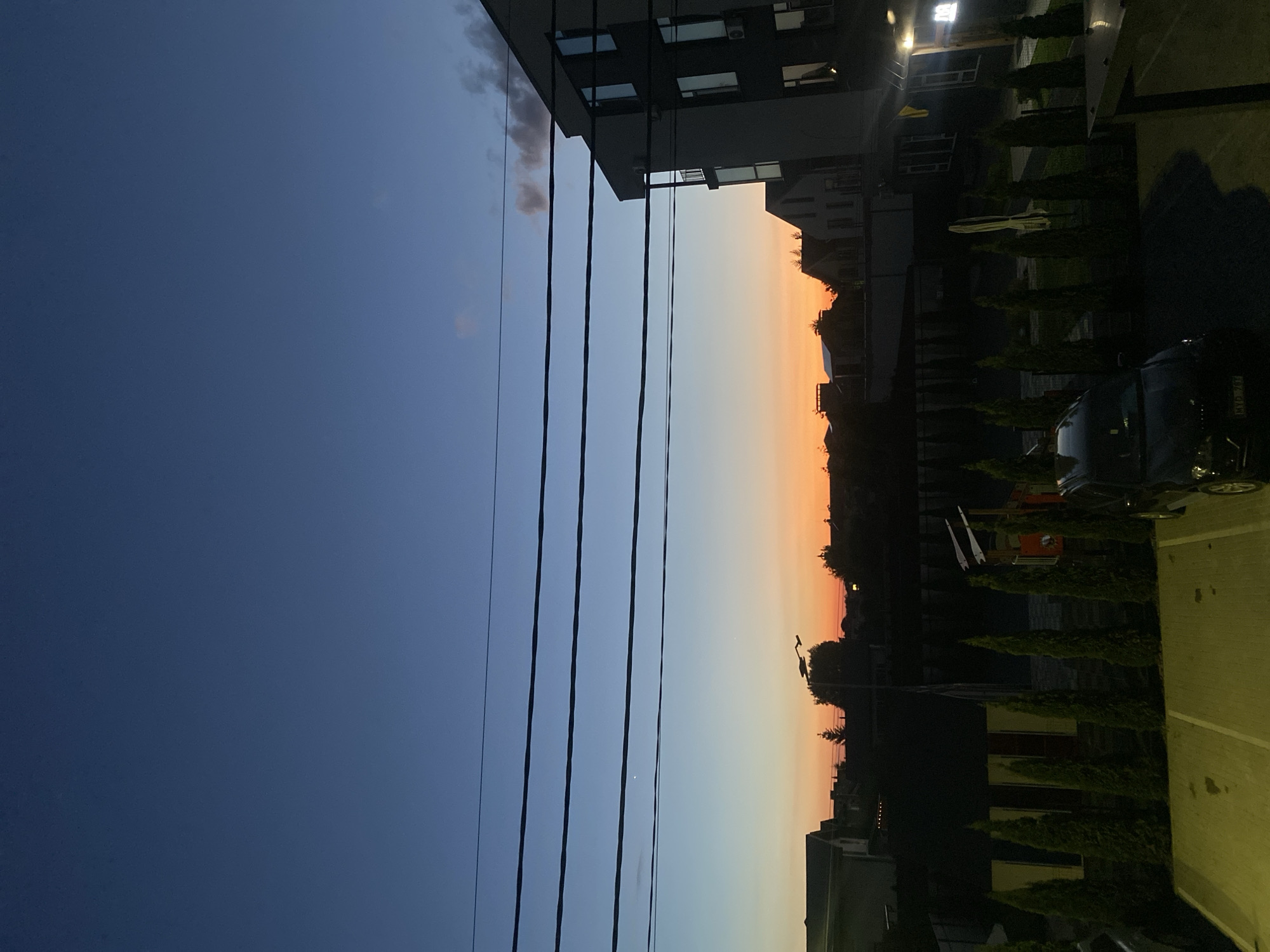 The image captures a twilight scene in Novi Petrivtsi, Ukraine, featuring a residential area under a gradient sky of deep blue transitioning to orange. In the foreground, a dark-colored SUV is parked in a designated parking lot with yellow markings. A row of manicured evergreen trees lines the edge of the parking lot. Behind the trees, several low-rise buildings and a playground structure are visible. In the mid-ground, a modern, multi-story building with a dark facade and numerous windows dominates the right side of the frame. Overhead, several power lines are stretched across the sky. The overall impression is one of a quiet evening in a suburban or semi-urban setting.