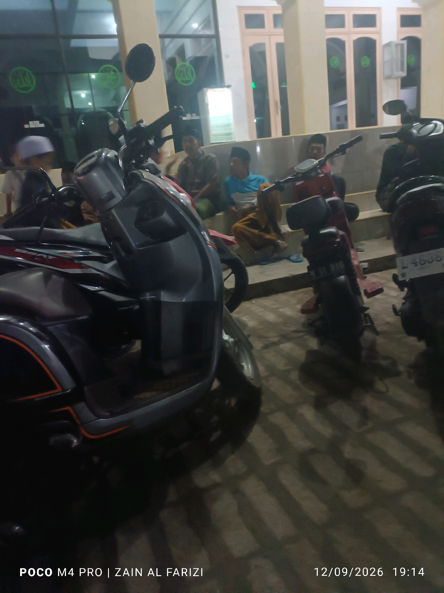 A low-angle, outdoor nighttime photograph captures a dimly lit scene with several motorcycles parked in front of a building with large windows. The building appears to be a mosque or similar religious establishment, as evidenced by the green Arabic calligraphy of "Allah" visible on the glass facade. In the foreground, a dark-colored scooter with orange accents is prominently featured, partially obscuring a portion of the scene. Behind it, a red scooter is visible, followed by another dark-colored motorcycle to the far right.
Seated on a ledge in the background are several individuals, appearing to be men, dressed in traditional attire. One individual in the middle wears a blue and white shirt and brown pants, while another to his left wears a patterned shirt and a peklong cap. To the far left, a person in a white peklong cap is partially visible. The ground is paved with rectangular stones, and shadows from the motorcycles and a light source are cast across it.
The timestamp "12/09/2026 19:14" is present in the bottom right corner, along with text identifying the camera as "POCO M4 PRO | ZAIN AL FARIZI." The context provided for the location as "Glagga, Indonesia" suggests this is a common gathering place for the local community.