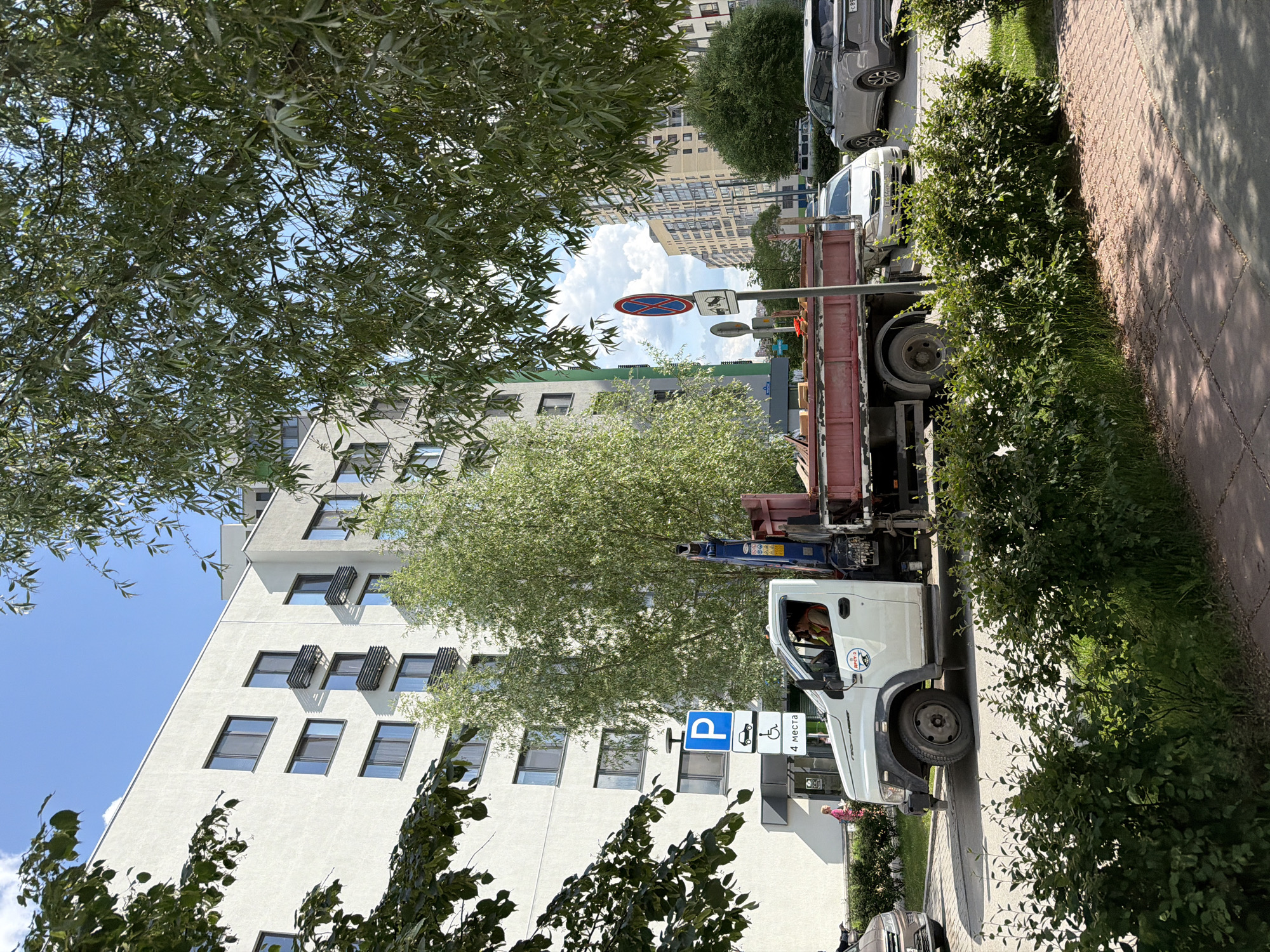 A white flatbed truck with a crane is parked on a paved road in front of a modern multi-story building. The truck has a red flatbed filled with construction materials. Two individuals are visible inside the truck's cab, with one appearing to be the driver. Traffic signs are posted on the roadside, including a parking sign with a blue 'P' and symbols indicating parking for cars and disabled individuals, as well as a "No Parking" sign. Lush green trees and bushes line the street, with a sidewalk made of brick pavers visible in the foreground. In the background, other residential buildings and a gray SUV are present. The sky is blue with some clouds. The location appears to be a residential area within the city of Tyumen, Russia, given the Russian text on the signs.