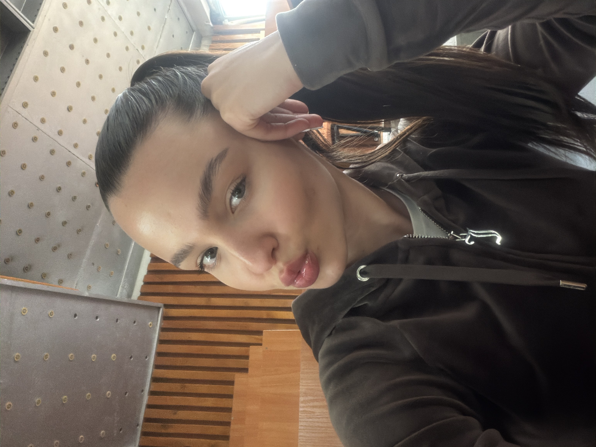 A young woman is captured in a headshot-style portrait, looking directly at the camera with a playful expression. Her head is tilted to the side, and her lips are pursed. She has dark hair pulled back, and her left hand is raised to her temple.
She is wearing a dark, possibly velour or velvet, hooded jacket with a visible silver zipper pull and silver aglets on the drawstring. A lighter-colored garment is seen underneath the jacket.
The background is divided, showing a light-colored wall or panel with a grid pattern of small, round, brass-colored studs or rivets on the left. On the right, there is a wooden surface characterized by horizontal slats, possibly part of a staircase or interior wall. The lighting appears to be natural indoor light, suggesting it is daytime. The overall mood is casual and lighthearted. No visible text is present. Given the location context, the setting is an indoor space in Sofia, Bulgaria.