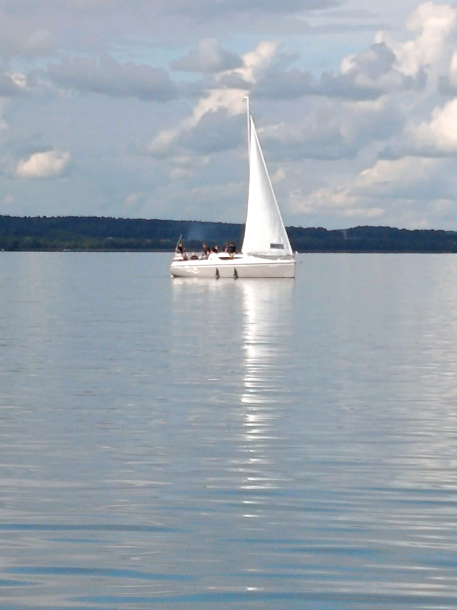 An outdoor, eye-level, medium shot captures a white sailboat with a white sail, navigating across a wide, calm body of water. The boat is in the middle ground of the image, slightly to the right of center, and appears to be a catamaran, with two hulls visible. A group of people, varying in number, are on the deck of the sailboat. The water is a pale blue and reflects the sky and the sailboat, creating streaks of light and shadow on its surface. In the background, a dark, tree-lined shore stretches horizontally across the frame, suggesting land. Above the land, a cloudy sky, filled with patches of white and gray clouds, dominates the upper portion of the image, with hints of blue sky peeking through. The lighting suggests it is daytime, possibly overcast or late afternoon. There is no indication of the specific location, city, or country.