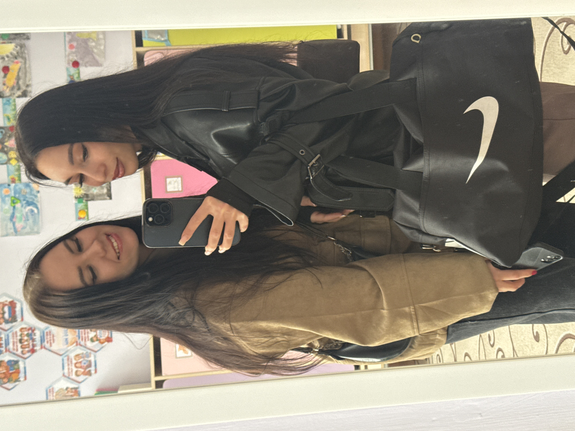 Two young women are captured in a mirror selfie within an indoor setting, likely in Chaikovsky, Russia. The image, oriented horizontally, shows their reflections in a large mirror.
One woman, positioned higher in the reflection, has long dark hair and is wearing a black leather-like jacket with buckles and straps. She is holding a dark smartphone, presumably an iPhone, to take the picture, and displays a pleasant smile. A black duffel or shoulder bag with a prominent white Nike swoosh logo is visible on her arm.
The second woman, positioned lower in the reflection, also has long dark hair and is wearing a light brown or tan jacket or coat. She is looking downwards with a soft, smiling expression. Her left hand is visible, with red nail polish on her thumb.
The background reflected in the mirror reveals a vibrant interior space. Colorful drawings and artworks, possibly children's creations, adorn a wall or surface behind them. Shelves or cubbies containing various objects, including what might be books or files, are also visible. A distinctive hexagonal pattern, possibly a decorative or educational chart with indistinct illustrations, can be seen on another wall surface. The women's jackets suggest cooler weather. Both subjects appear relaxed and happy, engaged in a casual moment together.