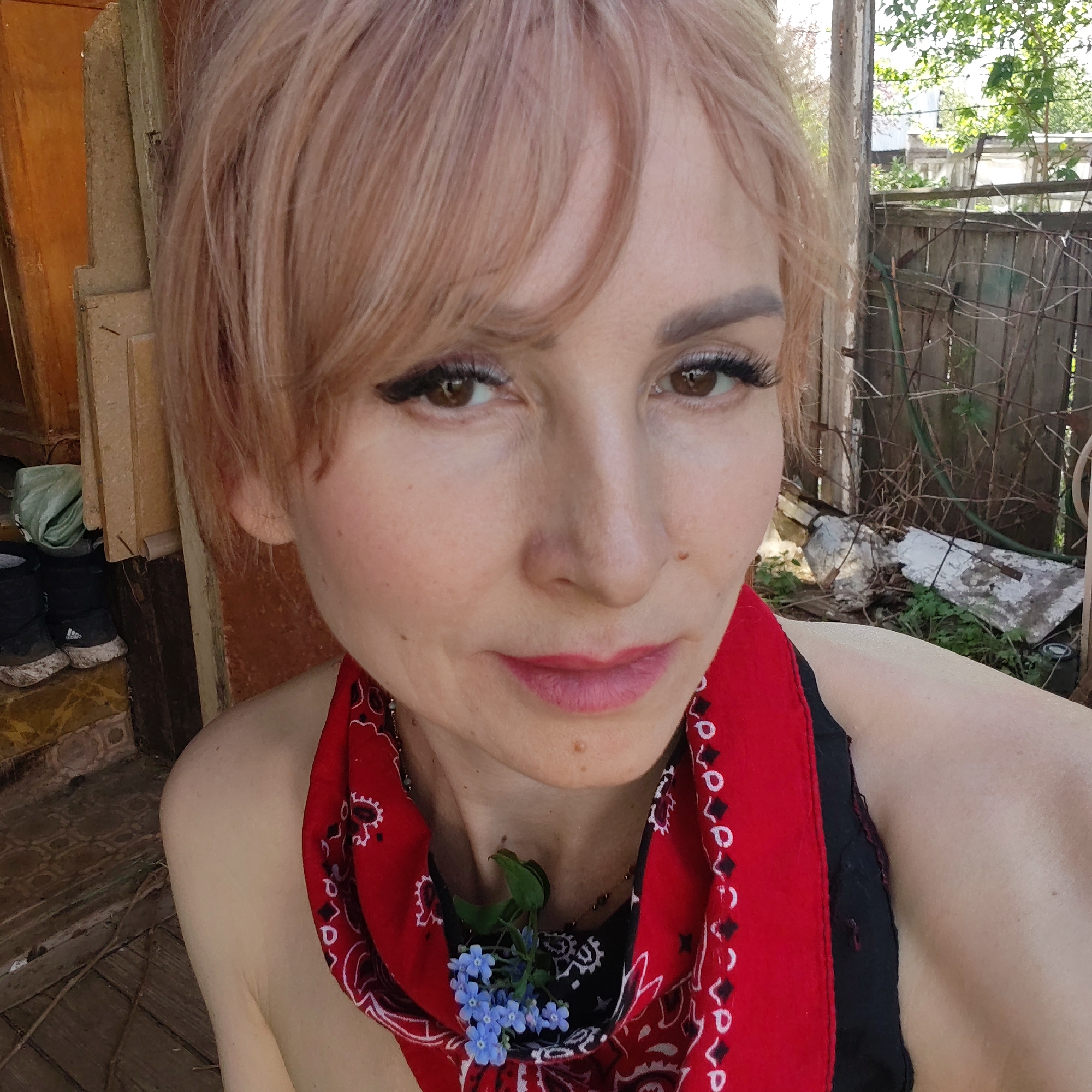 A close-up selfie shows a woman with short, light-pinkish blonde hair, fair skin, and brown eyes with visible eyeliner and mascara. She is wearing a red paisley-patterned bandana around her neck, which is partially tucked into a black top. A small cluster of blue flowers is tucked into the bandana near her chest.
She is standing outdoors, possibly on a porch. Behind her to the left is part of a weathered wooden structure, possibly a building or porch railing, with a pair of dark boots and a green bag visible on a tiled step. To the right, there is a view of a yard with a wooden fence and some vegetation. The location is Chaikovsky, Russia.