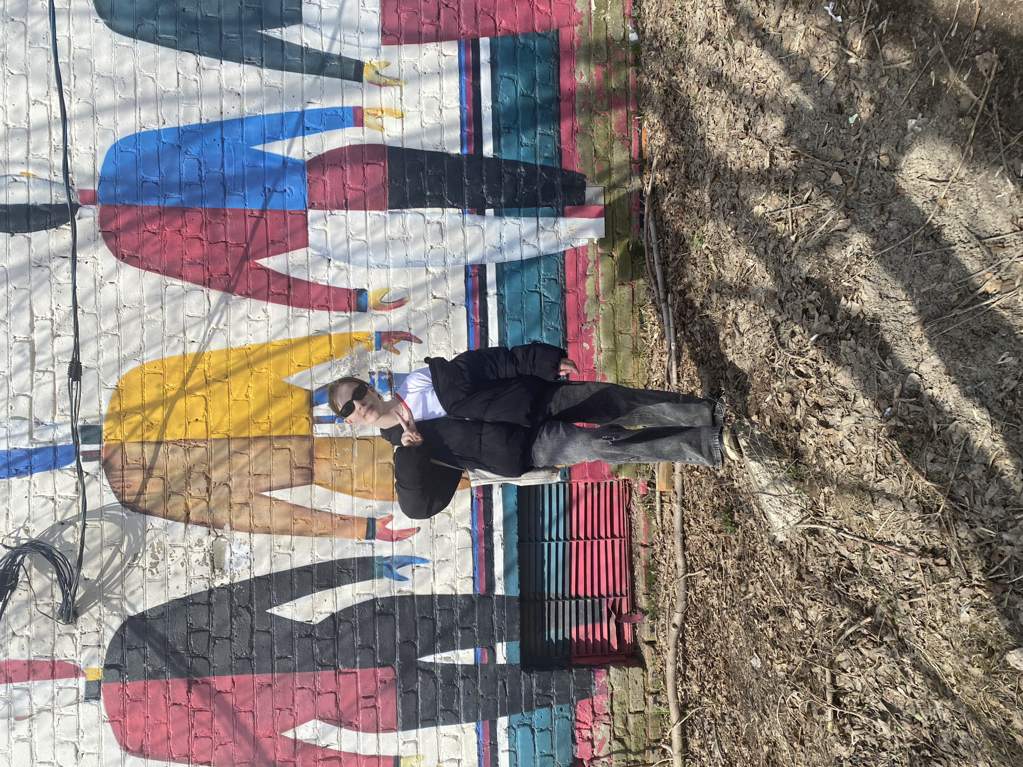 A young individual poses casually in front of a vibrant, abstract mural painted on a white brick wall in Chaikovsky, Russia. The person, positioned towards the right of the frame, wears a black puffer jacket, a white shirt, dark jeans, and sunglasses. They are making a peace sign with their left hand while looking at the camera. The mural depicts stylized, faceless figures or torsos in bold colors including red, blue, yellow, and black. A black cable runs vertically along the left edge of the wall.
To the right of the person, the ground is an unpaved area covered in dry leaves, dirt, and twigs, suggesting an unmaintained natural or semi-urban space. A metal grate, painted with red and blue stripes, is visible at the base of the wall near the person's feet. Bright sunlight illuminates the scene, casting long, distinct shadows across the ground and wall, indicating a clear, sunny daytime. No discernable text is visible.