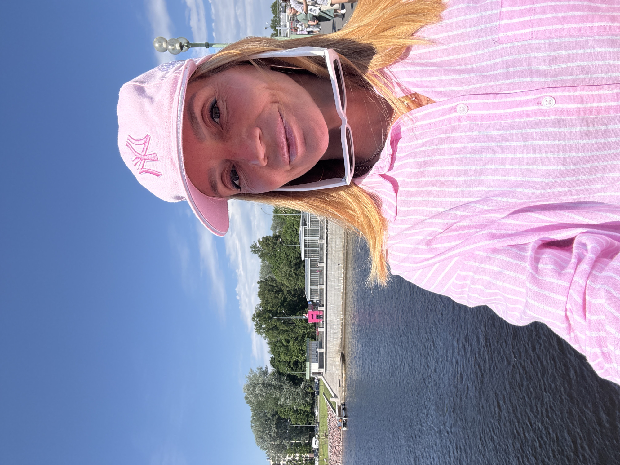 A woman with blonde hair and sunglasses perched on her head, wearing a pink striped button-down shirt and a pink New York Yankees baseball cap, takes a selfie on a waterfront promenade. The water in the foreground is dark blue. In the background, there is a promenade with a stone embankment, a walkway with railings, trees, and buildings. A large pink art installation is visible on the embankment. The sky is blue with scattered white clouds. The scene appears to be the Petrogradskaya Embankment in Saint Petersburg, Russia, near the "Bolshoy Dom" building, which is often adorned with art installations.