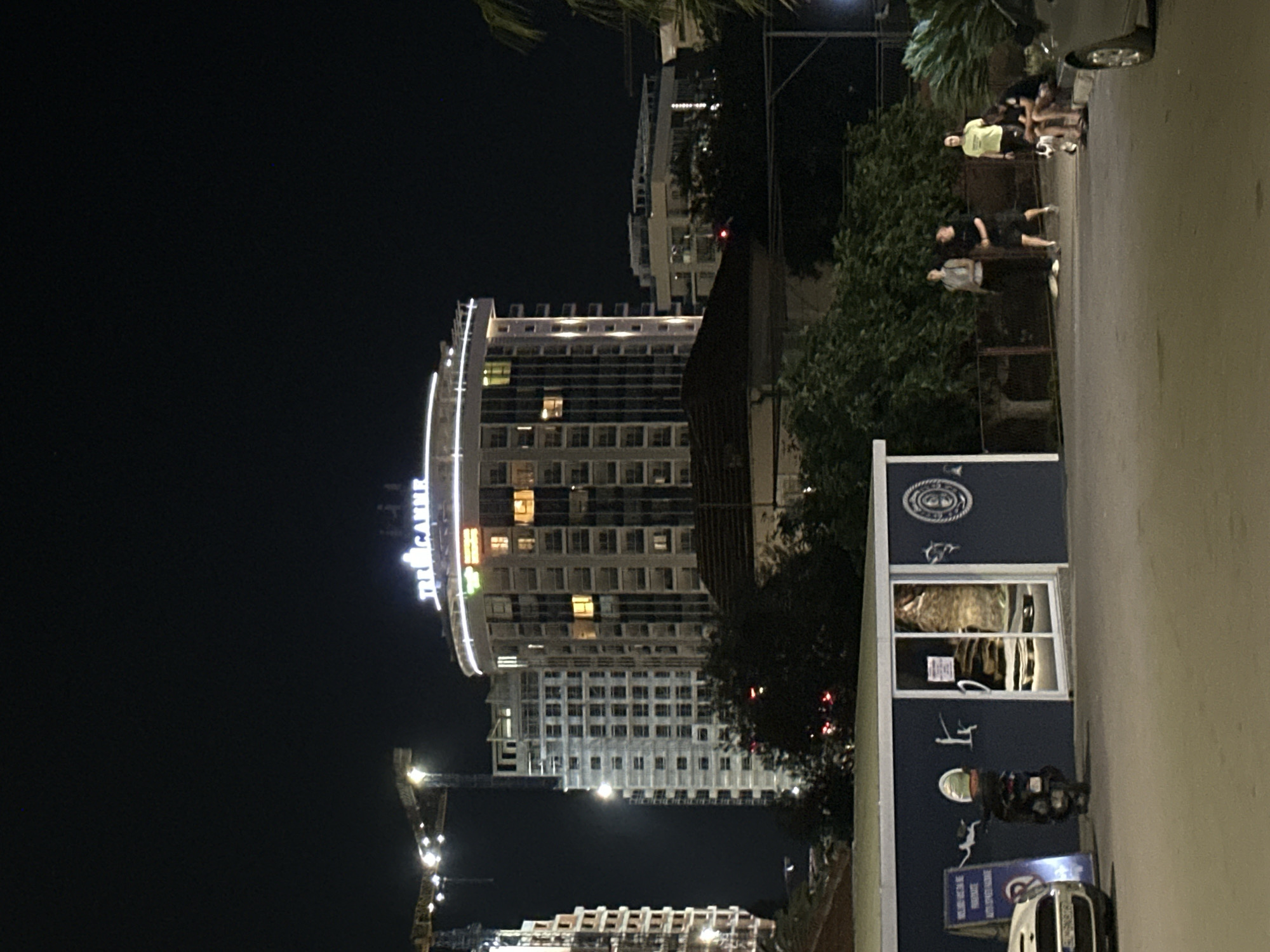 The image depicts a nighttime street scene in Budva, Montenegro. In the background is a tall building, illuminated with lights, bearing the name "TRE CANNE" in large, lit letters at the top. Adjacent to the building is a construction site with cranes also illuminated. The street is occupied by a group of people standing near a blue and white structure. A motorcycle is parked nearby. A car is parked in front of the structure. The scene is lit by streetlights and the ambient light from the buildings.