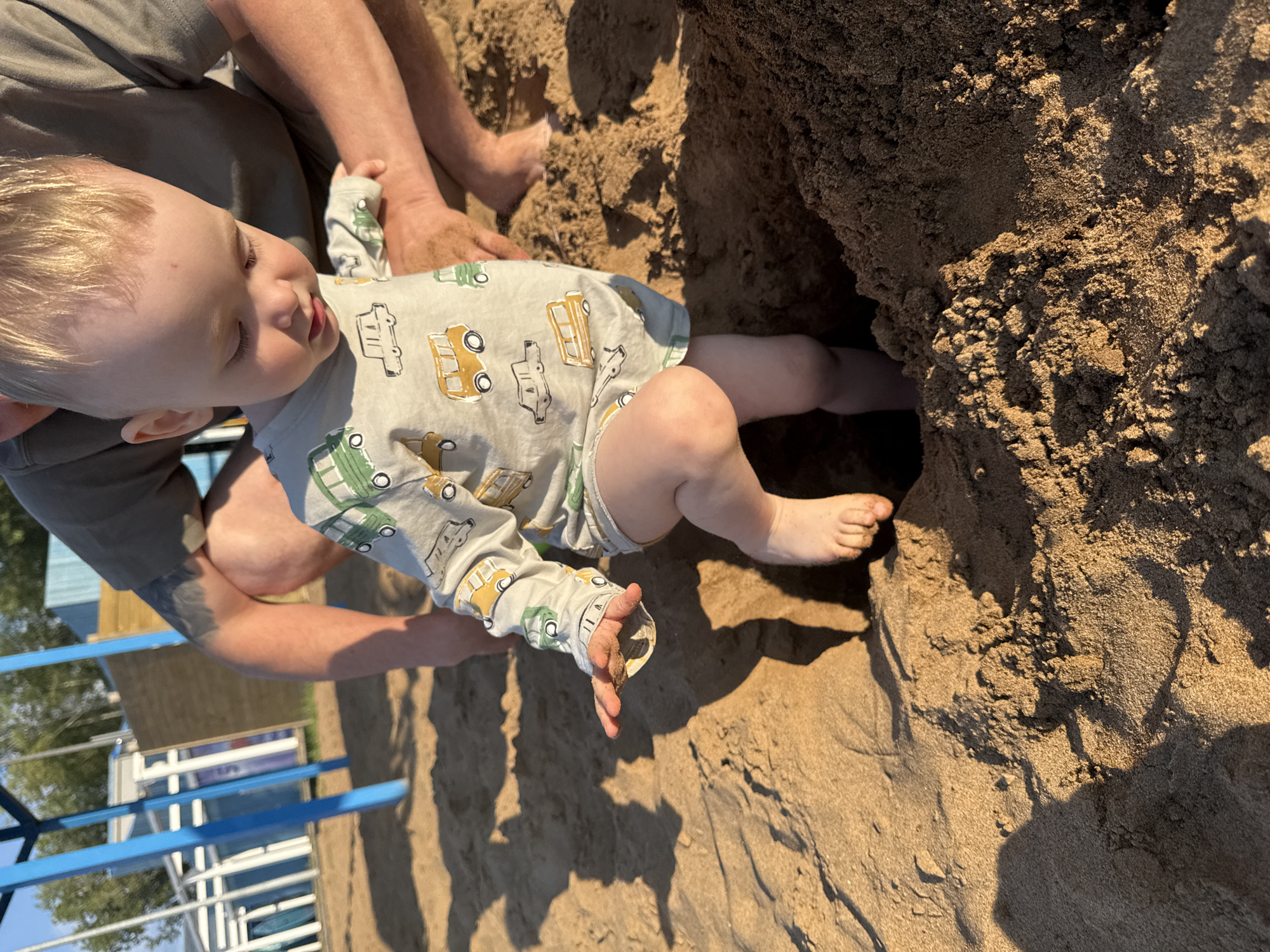 In this image, a baby with blonde hair is sitting on the sand, looking downwards. The baby is wearing a light-colored onesie with a pattern of yellow and green cars. The baby's left hand is holding a small amount of sand.
To the left of the baby, an adult's legs and torso are visible, suggesting the adult is sitting with the baby. The adult is wearing a grey t-shirt and their arm is resting near the baby.
The scene is set on a sandy area, possibly a beach or a sandbox, with a large mound of sand to the right of the baby. The lighting suggests it is daytime, likely late afternoon or early evening due to the warm, golden hue of the sunlight casting shadows on the sand. In the background, there are hints of a blue structure and trees, possibly indicating an outdoor recreational area. The overall mood appears relaxed and focused on the child's play.
The image is oriented sideways, with the top of the frame appearing on the left. No visible text is present in the image.