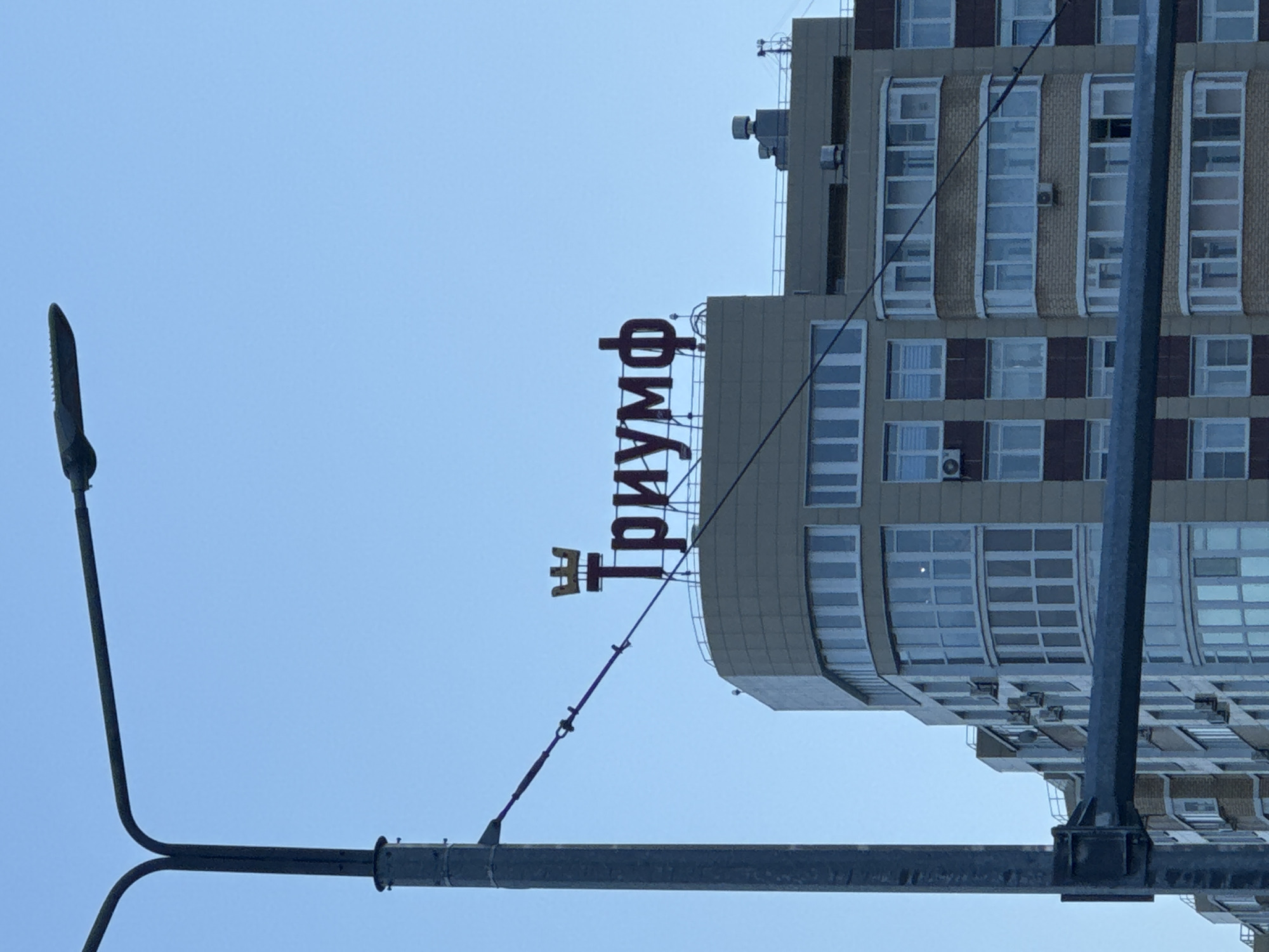 The image depicts a tall building in Perm, Russia, under a clear blue sky. The word "Триумф" (Triumph) is prominently displayed in red lettering on the building's upper facade. In the foreground, a streetlight pole extends upwards, partially obscuring the building. The building itself is multi-story, with a beige and brown facade and numerous windows. Several air conditioning units are visible, mounted on the building's exterior.