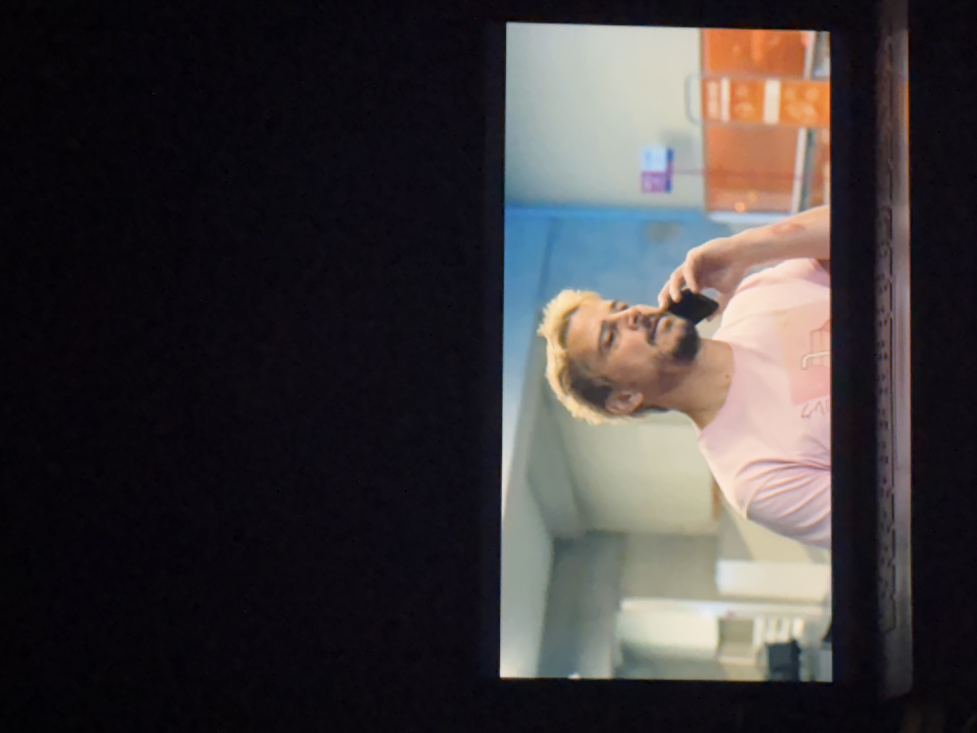 The image displays a rotated view of a screen, which shows a man engaged in a phone call, set against an indoor background. The screen occupies the right side of the frame, with a large dark, blank area to its left, and a dark border on its right.
The central subject is a man, depicted from the chest up and seen in profile, facing left within the screen's frame. He has light-colored (blonde or light brown) spiky hair and a neatly trimmed beard. He is wearing a light pink, short-sleeved t-shirt. His right arm is raised, with his hand holding a black smartphone to his ear, suggesting he is actively conversing. His expression appears neutral or slightly focused.
The background behind the man, within the screen's view, consists of a light-colored wall (possibly beige or light blue/grey) and indistinct structures. There are several orange rectangular elements, possibly shelves, signs, or parts of a partition, stacked vertically in the upper right. Further back, on the lower half, there appear to be additional light-colored surfaces or furniture. The lighting suggests a well-lit interior, likely during the daytime.
Due to the image's overall rotation (approximately 90 degrees clockwise), the man appears to be lying down on his left side, and the background elements are also oriented sideways. There is no clearly visible or legible text anywhere in the image. The scene takes place in Budva, Montenegro.