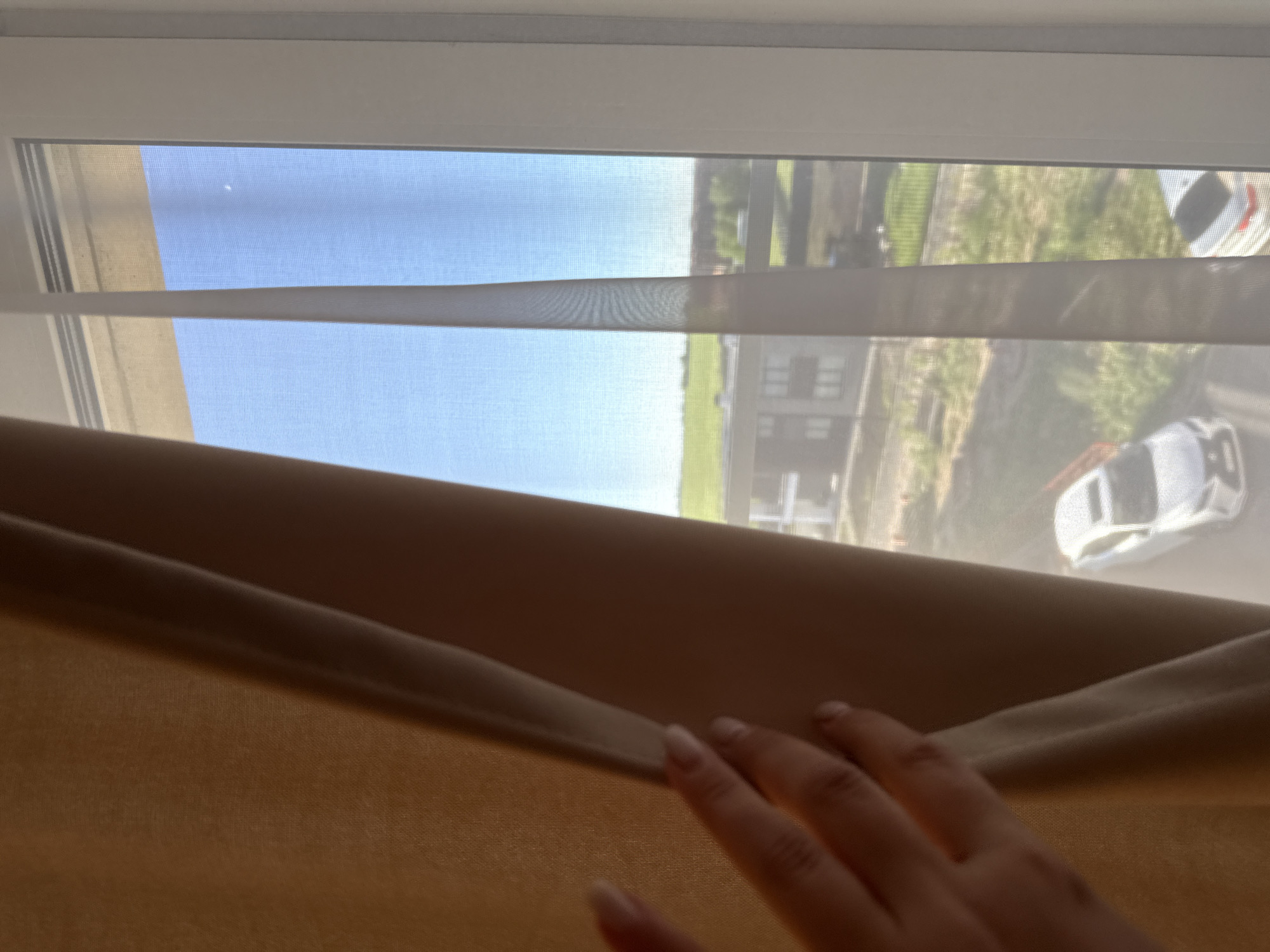 The image depicts a view from inside a room looking out through a window. In the foreground on the right, a person's hand with light-colored manicured nails is pulling back a textured, opaque, dark tan or brown curtain or blind. Behind this, a sheer, light-colored window shade with horizontal translucent bands is drawn across the window, allowing a partial view of the outside.
Through the window coverings, a clear, bright blue sky is visible in the upper portion, with a small, faint white dot, possibly the moon or a distant object, in the sky. Below the horizon, a green field or lawn stretches out. Further in the distance, modern-style buildings are visible, suggesting an urban or suburban environment. Several white cars are parked or moving on what appears to be a street or parking area.
The scene is set during the daytime under sunny, clear weather conditions, indicated by the bright natural light and blue sky. The activity shows someone subtly observing the outside world by peeking through the window coverings. The location context provided is Vologda, Russia. There is no visible text in the image.