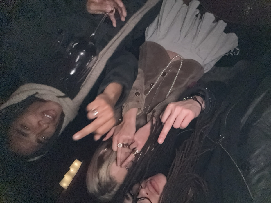 A gathering of three individuals in a dimly lit setting, likely indoors. Two people point downwards, each with a ring-adorned hand. One person is holding a wine glass. Attire includes a dress with ruffles, a brown velvet jacket, a light grey sweater, and a black leather jacket. The scene includes visible accessories such as a string of pearls and a beaded bracelet. One person has locs and glasses, another has blonde hair with dark roots.
