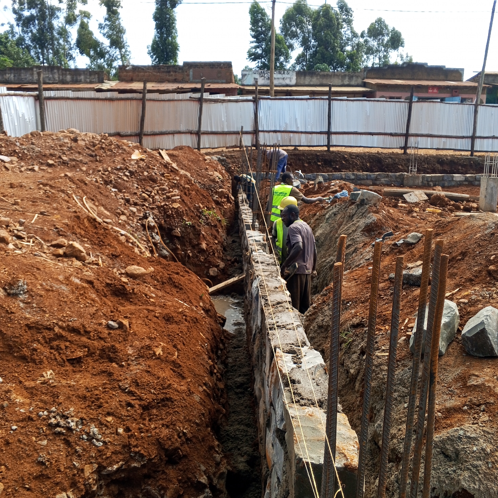 The image captures a construction site in Borabu Chitago ward, Kenya, during the daytime under clear skies. Several male construction workers are actively involved in building a stone foundation or retaining wall within a deep, excavated trench. The surrounding earth is a distinct red-brown soil, and some standing water is visible at the bottom of the trench alongside the newly laid stone wall.
One worker, wearing a yellow hard hat and a high-visibility vest with "1ST GROUP" printed on the back, stands near the wall. Another worker in a dark shirt and yellow hard hat is also visible closer to the camera, while a third in blue clothing works further in the background. Metal rebar protrudes from the ground, indicating future concrete elements of the structure.
In the background, behind a corrugated iron fence that encloses the site, there are several simple buildings and lush green trees. Visible text on one of the buildings faintly reads "UTLIMS CYBER & SALON". The scene depicts ongoing foundational work in what appears to be a developing area.