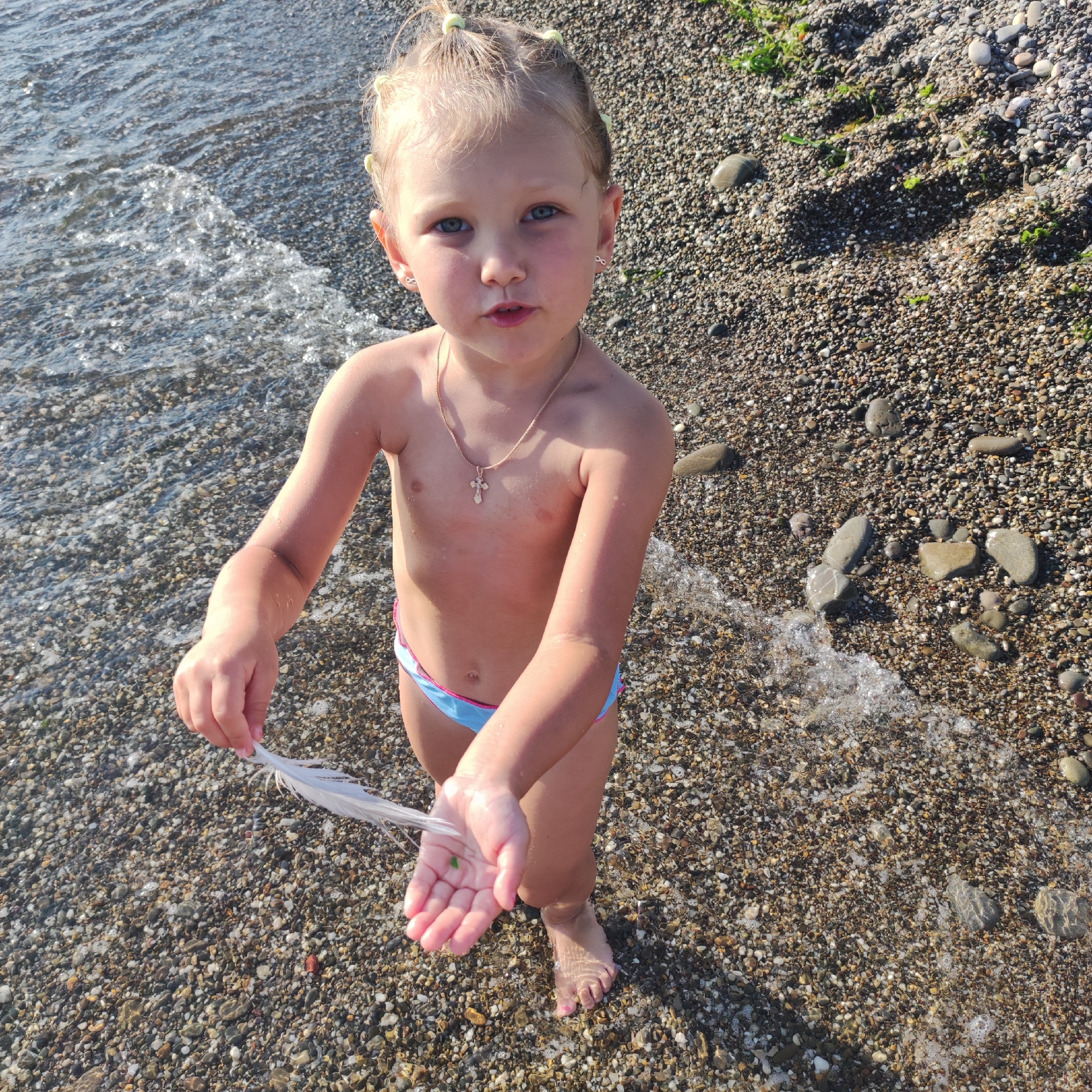 A young girl stands on a pebbled beach, facing forward and looking directly at the camera with a curious expression. She has blonde hair pulled up in several small ponytails, and is wearing a gold necklace with a cross pendant and small, silver, infinity-shaped earrings. Her torso is bare, and she wears blue and pink swim bottoms. In her outstretched hands, she holds a white feather. The water of the sea gently laps at her feet, which are submerged in the shallow water. The beach is covered in small, smooth pebbles of various colors, and a few pieces of green seaweed are visible. The lighting suggests it is daytime, likely sunny. The overall scene evokes a sense of childhood innocence and discovery on a beach vacation.