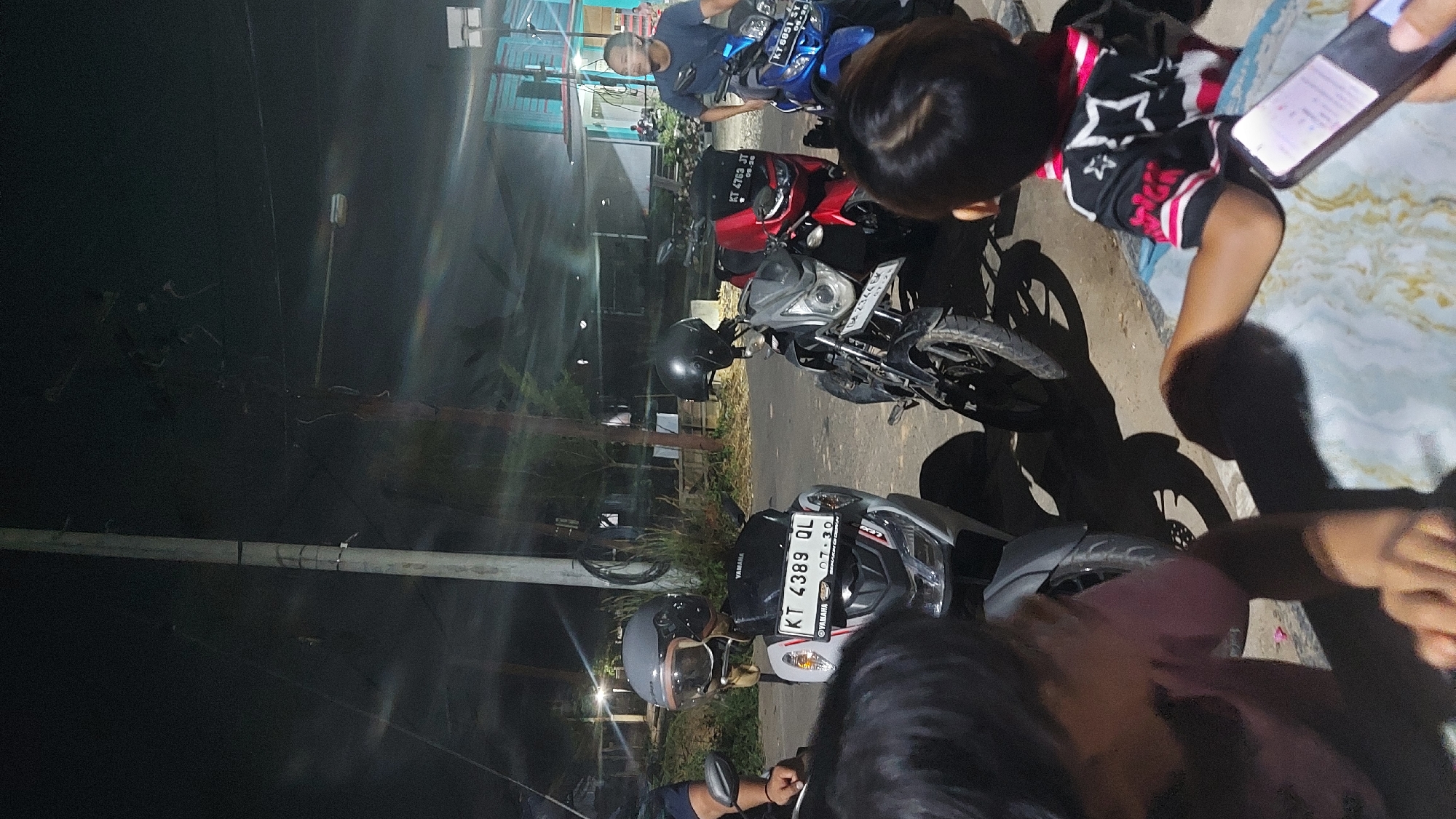 The image captures a nighttime scene in the city of Tanjung Redeb, Indonesia, with a group of people gathered around several parked motorcycles. A tall, light-colored utility pole stands prominently on the left side of the frame, with various wires and cables visible. In the foreground, two children are partially visible, one wearing a pink shirt and the other a black shirt with a white star pattern. A phone is being held by one of the children, displaying content on its screen.
Several motorcycles are parked in the middle ground. The closest is a light-colored Yamaha scooter with a grey helmet on its handlebars and a license plate reading "KT 4389 QL". To its right is a black motorcycle with a black helmet on its handlebars and a license plate that is partially obscured. Further to the right is a red motorcycle with a license plate "KT 4763 JT", and behind it, a blue motorcycle with a license plate "KT 6851 SY".
A man wearing a blue t-shirt is seated on the blue motorcycle, looking towards the right of the frame. In the background, there are structures that appear to be buildings, with some lights illuminating the area. The ground is paved with asphalt or concrete. The overall atmosphere suggests a casual gathering, possibly at a roadside stop or a communal area in Tanjung Redeb.