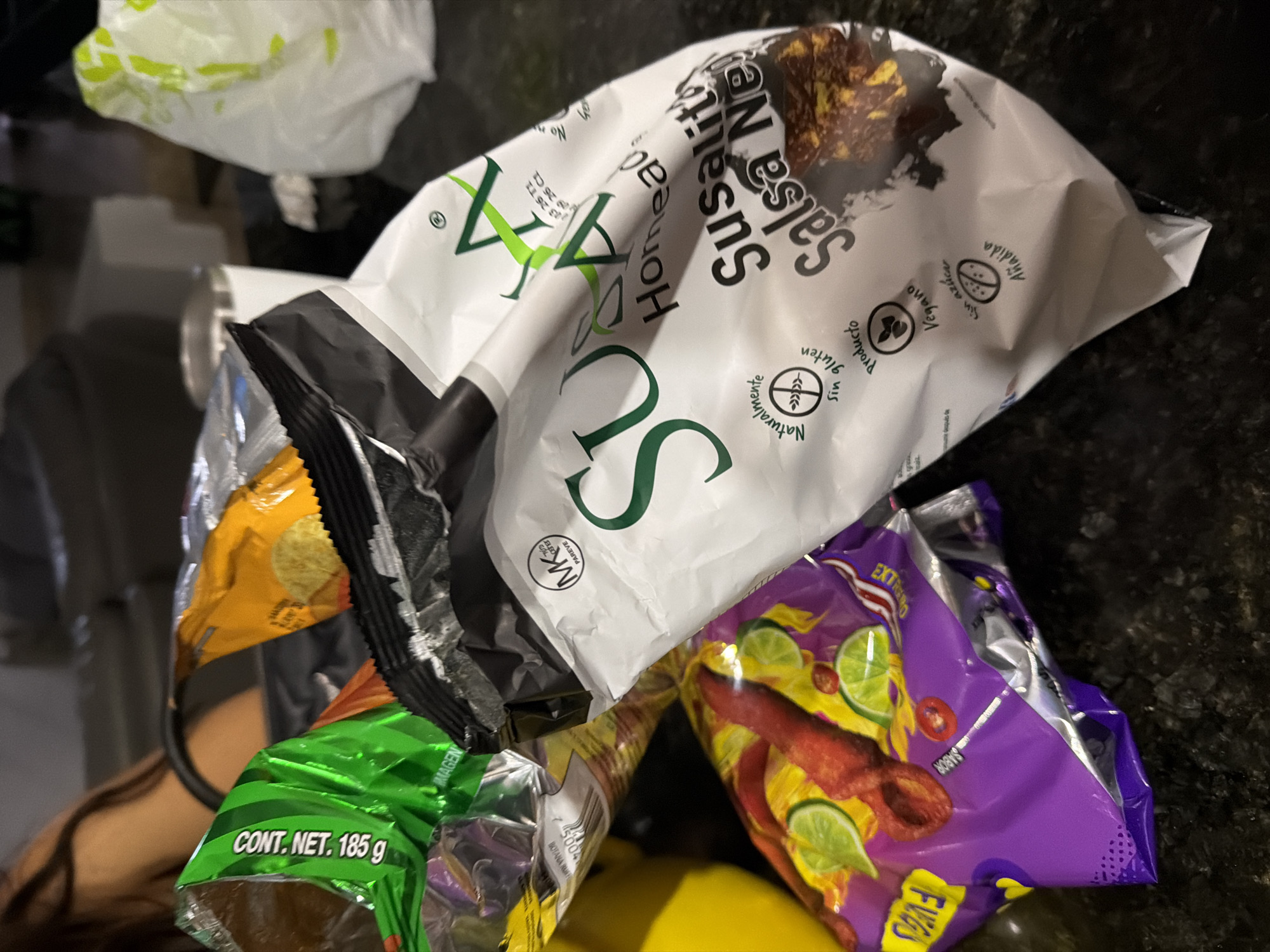 The image shows a pile of opened snack bags on a dark countertop. Visible brands and types of snacks include "Susalitas Salsa Negra", "Takis Fuego" (specifically, "EXTREMO" flavor), and another green bag labeled "CONT. NET. 185 g" of an unidentifiable snack. The scene appears to be indoors. The OCR suggests the presence of the "MK PAREVE" symbol, and information suggesting the product is from Mexico, specifically referring to ingredients and packaging. The image does not contain enough information to determine its exact location in Santa Clara Ocoyucan, Mexico.