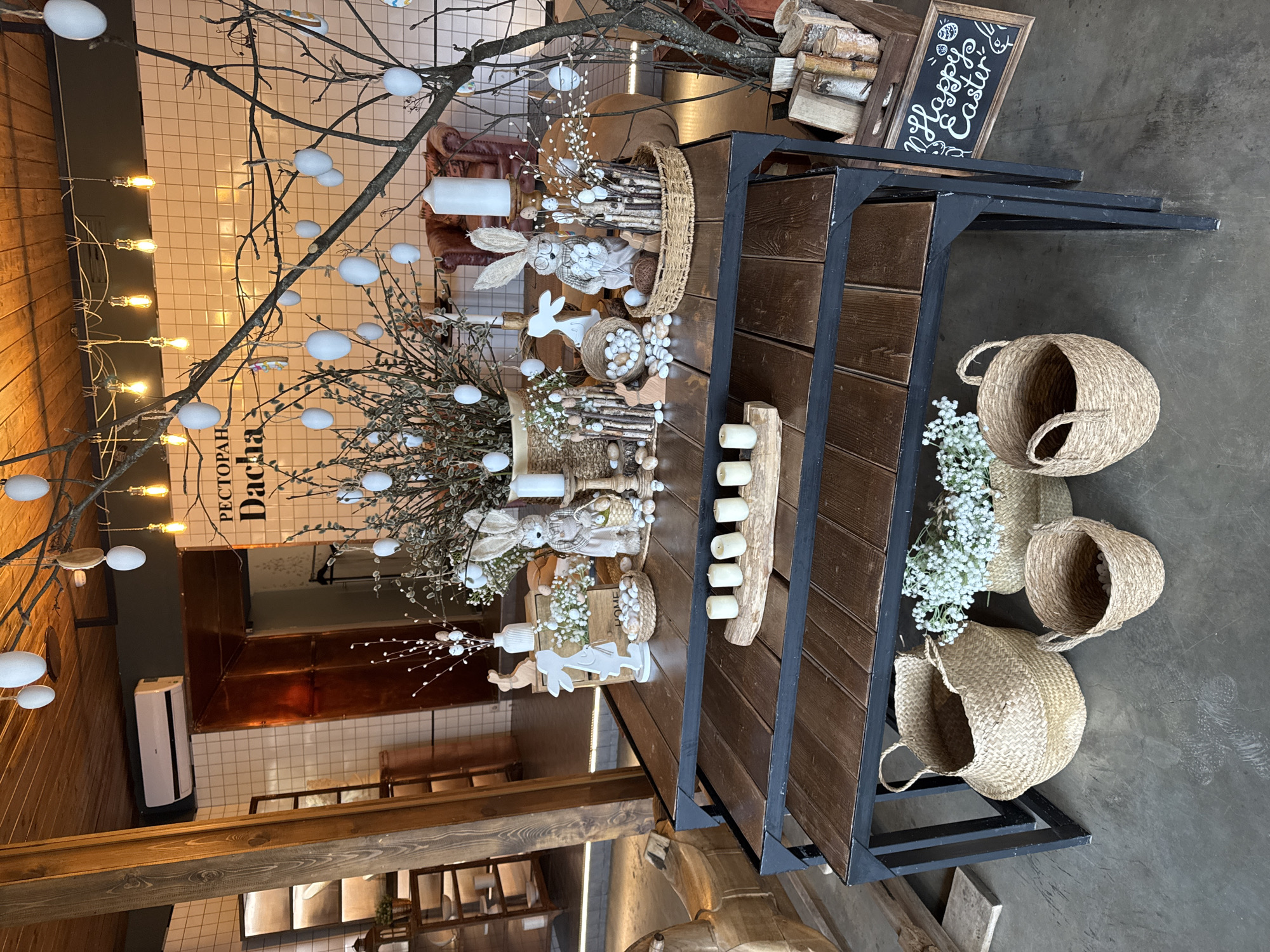 Captured from an elevated perspective, the image presents an elaborate Easter-themed display at a restaurant named "Dacha," the location of which remains undisclosed. The setting is defined by a harmonious blend of rustic aesthetics and festive arrangements.
At the forefront, a multi-tiered wooden table, reinforced with black metal supports, acts as the focal point. The table is adorned with baskets containing decorative Easter eggs, crafted wooden bunnies, and floral arrangements featuring white baby's breath. These elements are interspersed with candles, adding to the scene's ambient charm. Above, a tree branch, embellished with artificial white eggs, casts a whimsical shadow, while a string of soft-glowing lights further enhances the atmosphere.
To the left, glimpses of the restaurant's interior reveal wooden paneling and tiled walls, evoking a sense of rustic elegance. The background features additional seating arrangements and decorative elements, contributing to the overall festive ambiance.
The floor beneath the table is bare and utilitarian, contrasting with the decorative abundance above. The presence of baskets and flowers spilling onto the floor adds a touch of unstudied spontaneity to the scene.