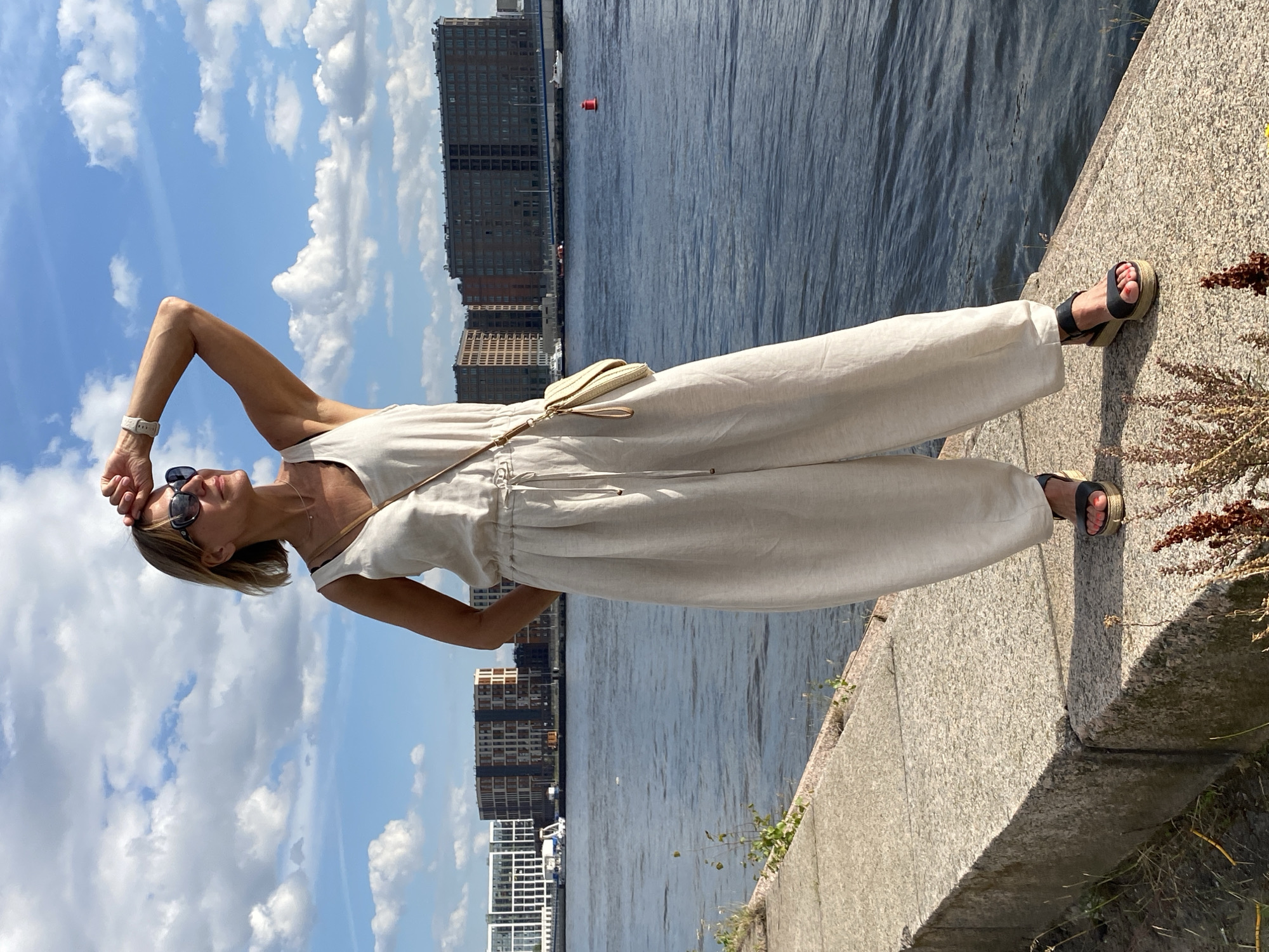 A woman stands on a granite embankment overlooking a body of water in Saint Petersburg, Russia. She is wearing a light-colored sleeveless jumpsuit, sunglasses, and dark-strapped platform sandals. Her right arm is raised, shielding her eyes from the sun as she looks out towards the horizon. A small, light-colored crossbody bag is slung over her left shoulder. In the background, several multi-story buildings line the opposite bank of the water, and the sky is partly cloudy. The water appears calm, with a few boats visible in the distance.