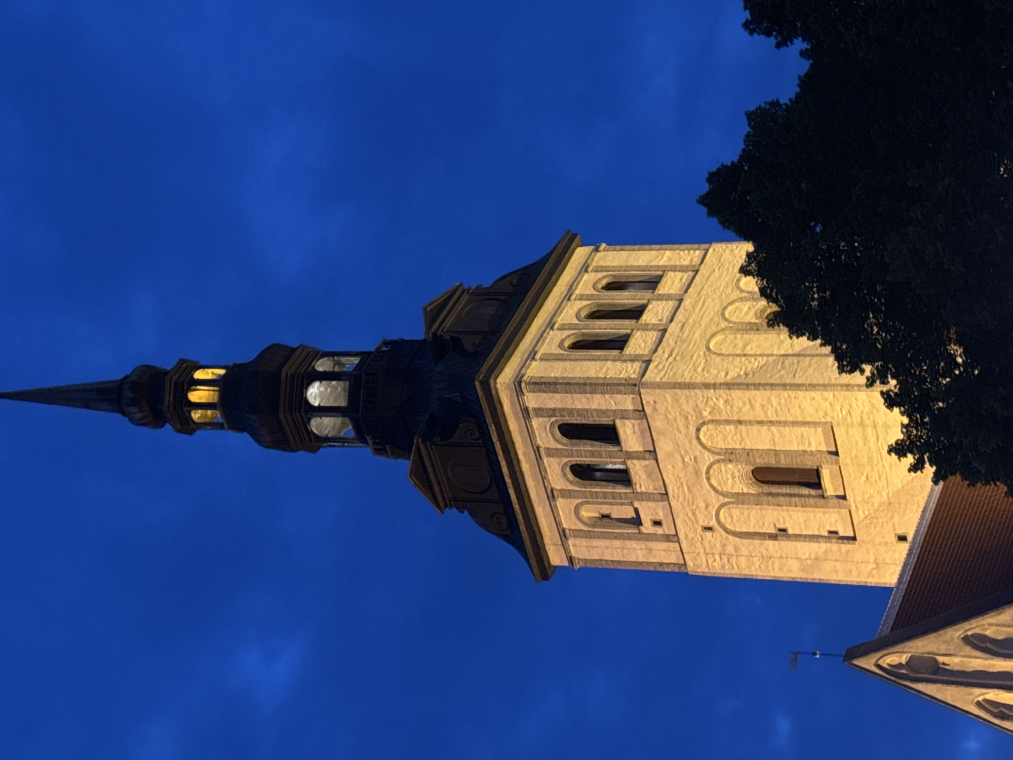The image shows the brightly illuminated tower of St. Olaf's Church (Oleviste kirik) in Tallinn, Estonia, against a deep blue twilight sky. The tower, constructed of light-colored stone, features multiple levels with arched windows. The spire at the top is dark and tapering, with illuminated sections visible within its upper levels. Dark silhouettes of trees are visible in the foreground on the right side of the frame, partially obscuring the lower portion of the tower and a section of a red-tiled roof to the left. A small flag is visible on a pointed architectural element above the roof. The overall impression is of a historic landmark at dusk, with artificial lights highlighting its architecture.