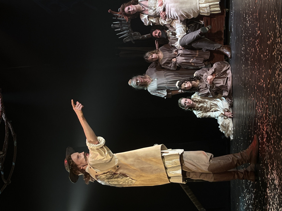 This image depicts a stage performance occurring at a theater or cultural center in Novoshakhtinsk, Russia. Seven individuals in period-style costumes are illuminated by spotlights on a dark stage, which features scattered light-colored fragments on its surface. On the far left, a male actor with a mustache and long, dark hair, dressed in a light-colored shirt, a tan vest with a metallic design, tan trousers, and dark brown lace-up boots, stands in profile. He extends his right arm directly forward, pointing with his index finger. To his right, a group of six other individuals is arranged. An older woman with light hair and a pale face, wearing a long grey dress and necklace, stands centrally. Beside her, a young girl in a dark brown dress stands with her gaze directed forward-left. In the front row, two young women are seated on the floor, both wearing long dresses (one cream, one dark brown) and looking forward-left. On the right, a man with a full dark beard and a woman with elaborate reddish headwear are seated on a dark wooden chest-like prop. Both are dressed in light-colored period attire and display serious expressions. Overhead, an abstract, dark, skeletal-like structure is partially visible, and a similar jagged prop serves as a backdrop behind the seated group on the right.