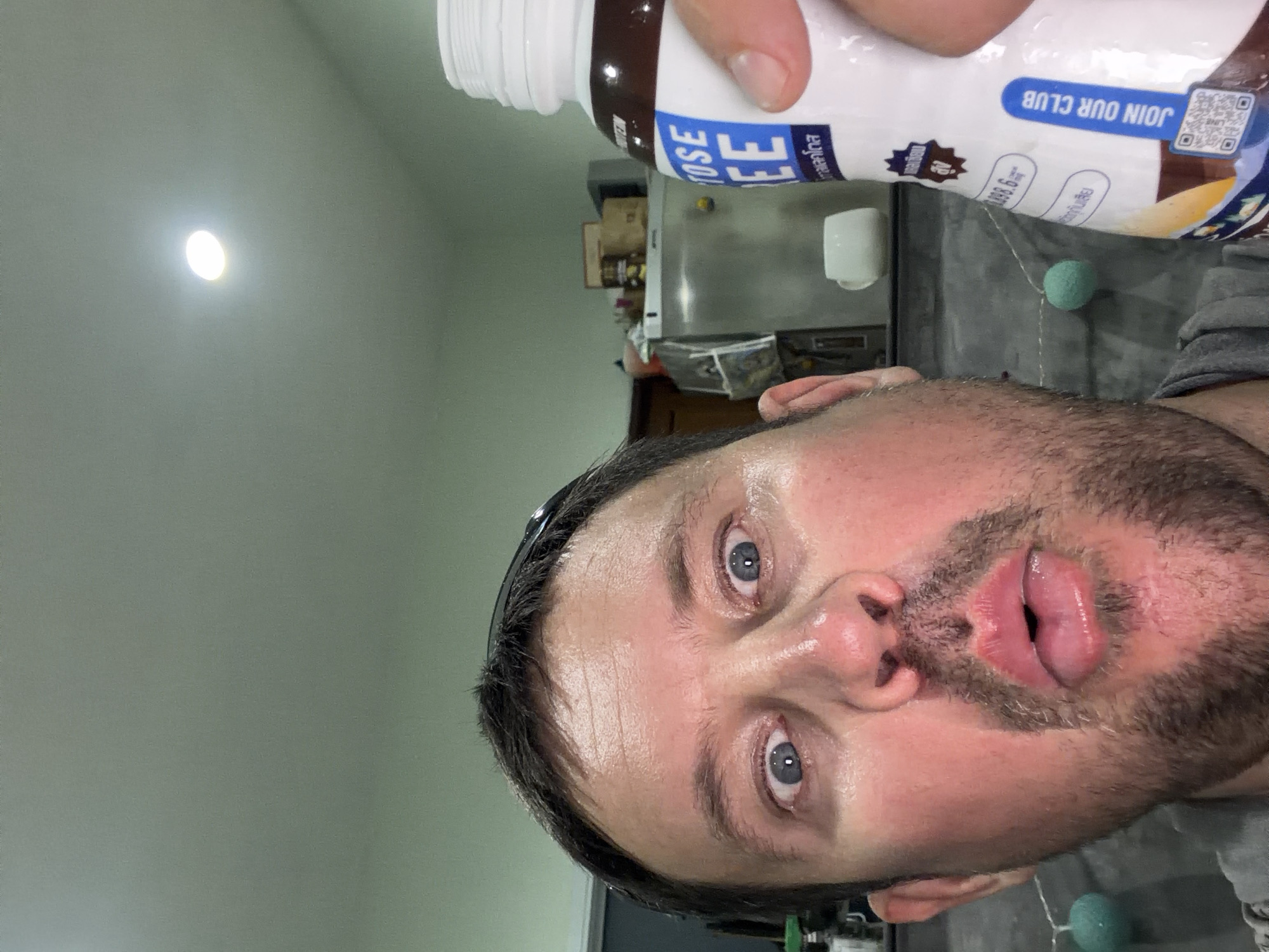 In Baan Mae Nam, Thailand, a close-up, selfie-style photograph captures a Caucasian man in his 30s or 40s, holding a bottle of "LACTOSE FREE" chocolate milk. The man has fair skin, blue eyes, and short brown hair, with stubble on his chin and upper lip. His eyes are wide open and his mouth is slightly ajar, creating a surprised or playful expression. He is wearing a gray t-shirt.
The background suggests an indoor setting, possibly a kitchen or pantry area, with a stainless steel appliance and a white mug visible on a shelf. The lighting is warm, with a ceiling light illuminating the scene. The man is holding the bottle of chocolate milk in his right hand, which is partially visible in the frame. The bottle has a white label with blue text, and a QR code is present on the lower part of the label. The man is positioned in the foreground, taking up most of the frame. The image has a casual, informal feel, typical of a personal snapshot.