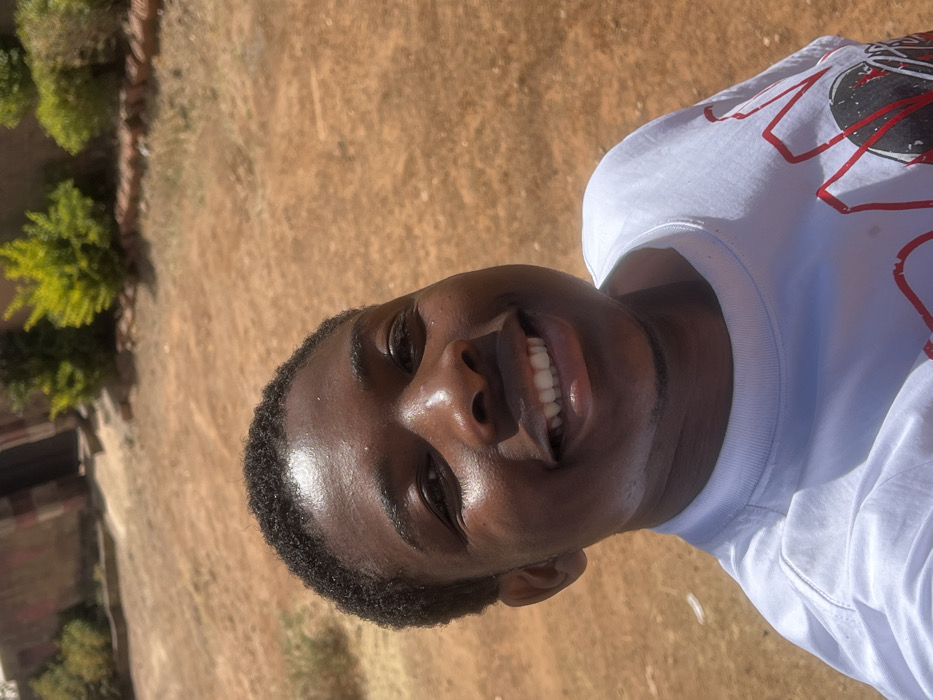 An outdoor image, taken in Kirana, Nigeria, captures a young individual with dark skin and short, dark, curly hair, positioned in a close-up, slightly upward-angled view. The individual is smiling broadly towards the camera, revealing white teeth, and wears a white short-sleeved t-shirt with a partially visible red and black graphic design on the chest. The background features dry, reddish-brown earth with sparse, desiccated vegetation patches. Behind the individual, in the mid-ground, are several small green ornamental shrubs. To the upper left, a building constructed of reddish-brown material with darker vertical banding and a partially visible dark, recessed doorway is present. The scene is brightly illuminated by natural daylight.