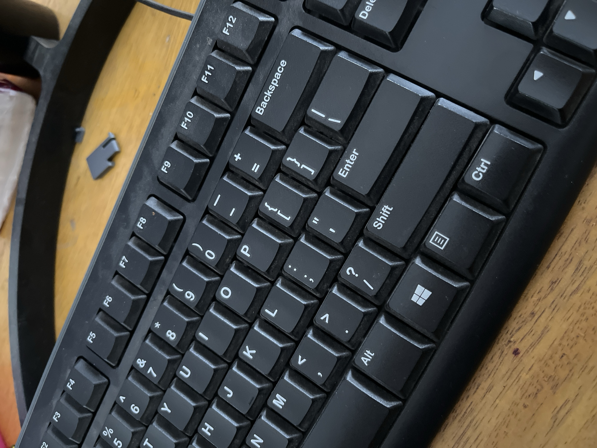 The image shows a close-up, high-angle shot of a black computer keyboard resting on a wooden surface. The keyboard is angled, with the top edge towards the viewer's upper right. Several keys are clearly visible, including function keys F2 through F12, number keys, letter keys, and modifier keys such as Alt, Shift, Ctrl, and the Windows key. Some keys have symbols printed on them, such as percentage, ampersand, asterisk, and punctuation marks. A small, gray plastic piece, possibly a keycap or a part of a computer accessory, is visible on the wooden surface to the left of the keyboard. The background is out of focus, showing a dark, curved object, likely part of a monitor stand or a similar piece of computer equipment. The lighting suggests an indoor environment. There are no people or animals in the image. The scene is static and appears to be a detail shot of computer hardware. There is no information in the image to suggest the location is the city of Kedawung, Indonesia.