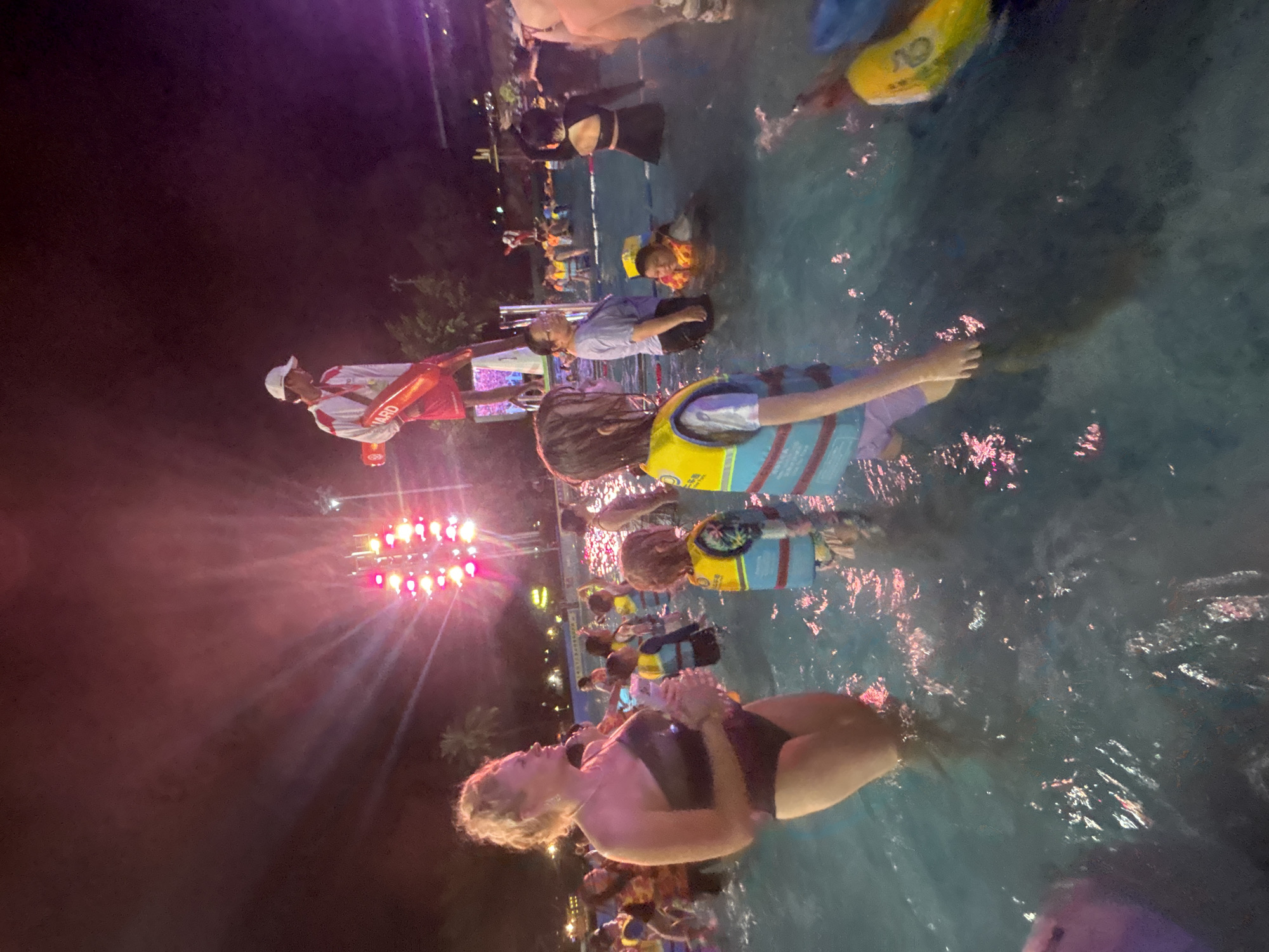An outdoor nighttime scene at a water park, likely in Panyu District, China, shows a crowd of people wading in a pool illuminated by bright pink and purple lights from a stage or light installation. A lifeguard, wearing a white cap, red shorts, and a white shirt with red trim and the word "GUARD" on it, stands elevated on a platform holding a red rescue buoy. Several children and adults are in the water. Some children are wearing life vests in yellow and blue. A woman in a dark one-piece swimsuit stands in the foreground, looking to her left. The water reflects the colorful lights, creating a vibrant and lively atmosphere.
