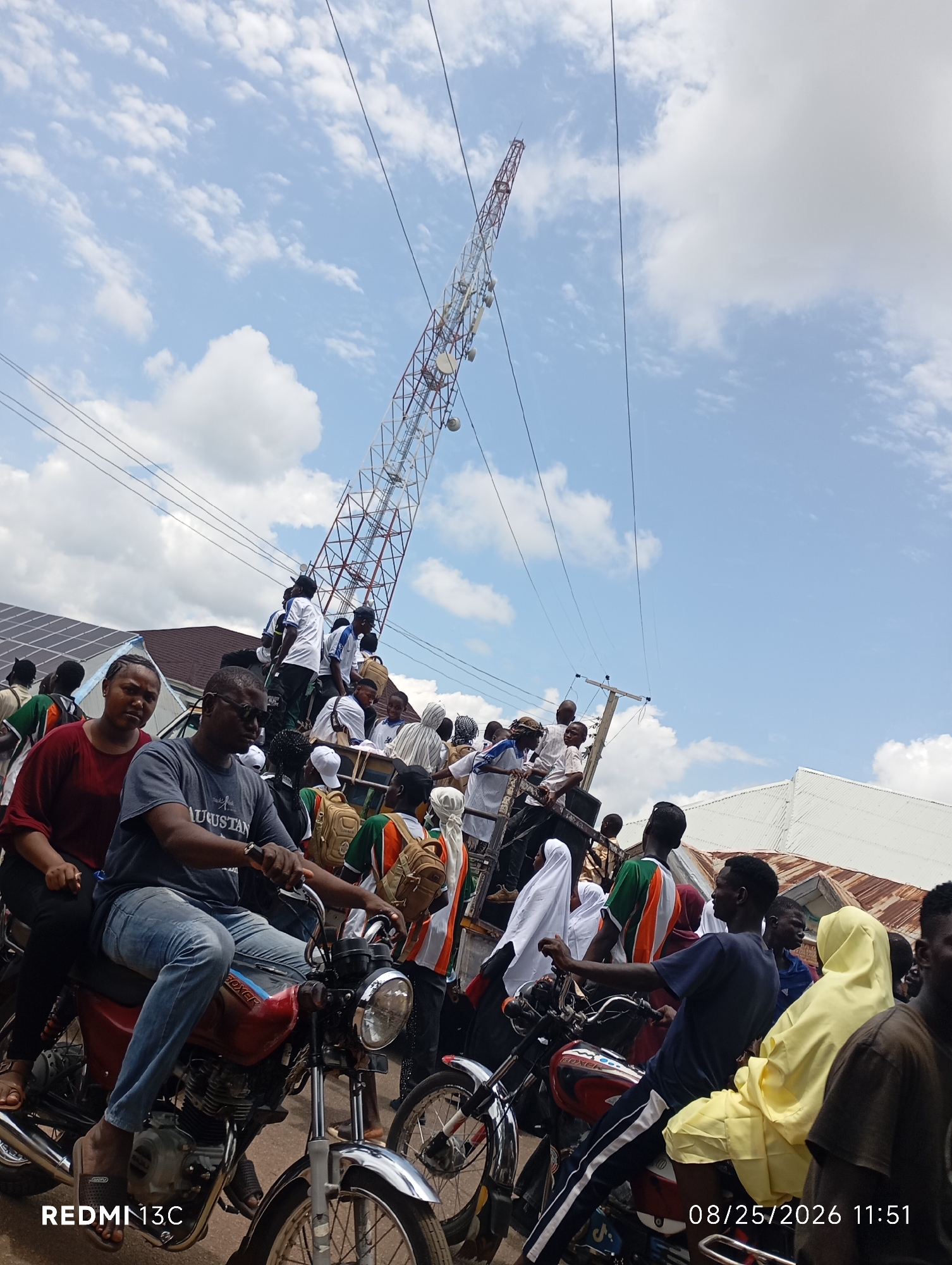 In Shendam, Nigeria, on August 25, 2026, at 11:51 AM, a crowd of people gathers under a tall telecommunications tower. The sky is partly cloudy. Many individuals are perched on a vehicle, possibly a truck, with some standing on motorcycles and a flatbed trailer. The people are dressed in casual clothing, with some wearing white shirts with blue accents and others in green, orange, and white attire, suggesting a possible team or group affiliation. Several individuals are carrying backpacks. In the foreground, two motorcycles are visible. The person on the left motorcycle, wearing a gray t-shirt with the word "AUGUSTAN" printed on it, is operating the vehicle. The person on the right motorcycle, wearing a blue t-shirt and dark pants with white stripes, is also operating the vehicle. The scene appears to be a gathering or procession, with people moving through the city. In the background, solar panels are visible on a building, and other structures line the street.