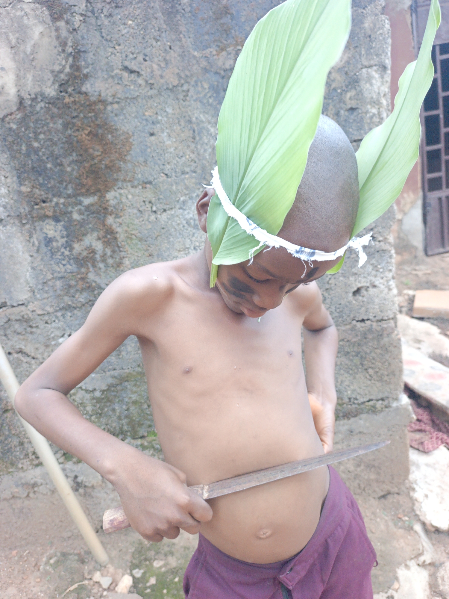 A shirtless, dark-skinned boy stands outdoors in Akwanga, Nigeria. He has a shaved head adorned with large green leaves secured by a white headband. Dark smudges are on his face, suggestive of face paint. He is holding a large, dull-looking knife horizontally against his abdomen. He wears maroon shorts. Behind him, a weathered grey wall is visible. To the right is a red object and a dark blue metal window frame. A light-colored pole rests against the wall to his left. The ground is dirt and sparse vegetation.