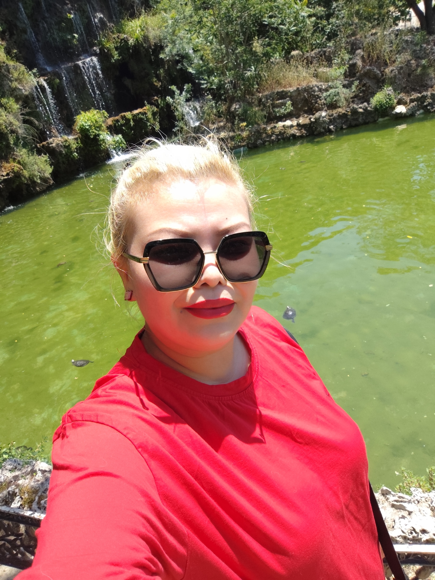 A woman with blonde hair pulled back and wearing large, dark-rimmed sunglasses and a bright red top is taking a selfie outdoors. She has bright red lipstick on. In the background, a green pond or lake is visible, with several turtles swimming in it. To the left of the pond, a multi-tiered waterfall cascades down a rocky, vegetated cliff face. Trees and vegetation line the top of the cliff. The overall setting appears to be a park or naturalistic garden. This location is likely the Polonezköy Nature Park, which is situated within the Sarıyer district of Istanbul, Turkey.