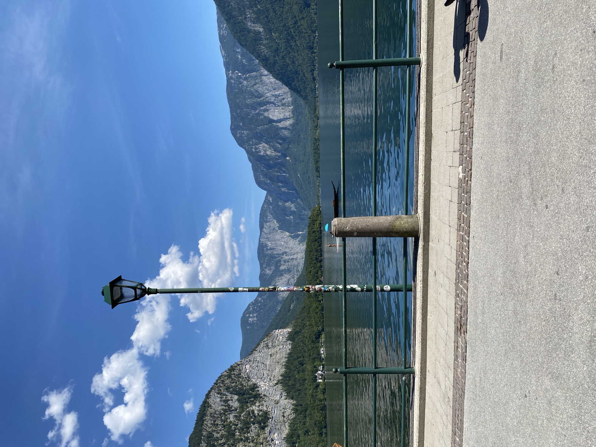 The image shows a scenic view of Hallstatt, Austria, on a sunny day. In the foreground, a paved walkway runs alongside a green metal railing that borders a calm, dark blue lake. A vintage-style lamppost, adorned with stickers, stands prominently on the walkway. Beyond the railing, the serene lake reflects the surrounding mountains. A lone gondola-like boat is visible on the water. In the distance, majestic mountains covered in lush green trees rise sharply, their rugged cliffs illuminated by the sunlight. The sky above is a clear, vibrant blue, dotted with a few fluffy white clouds. A small portion of the road is visible in the bottom of the frame, with the shadow of a person on a bicycle or scooter on the right.