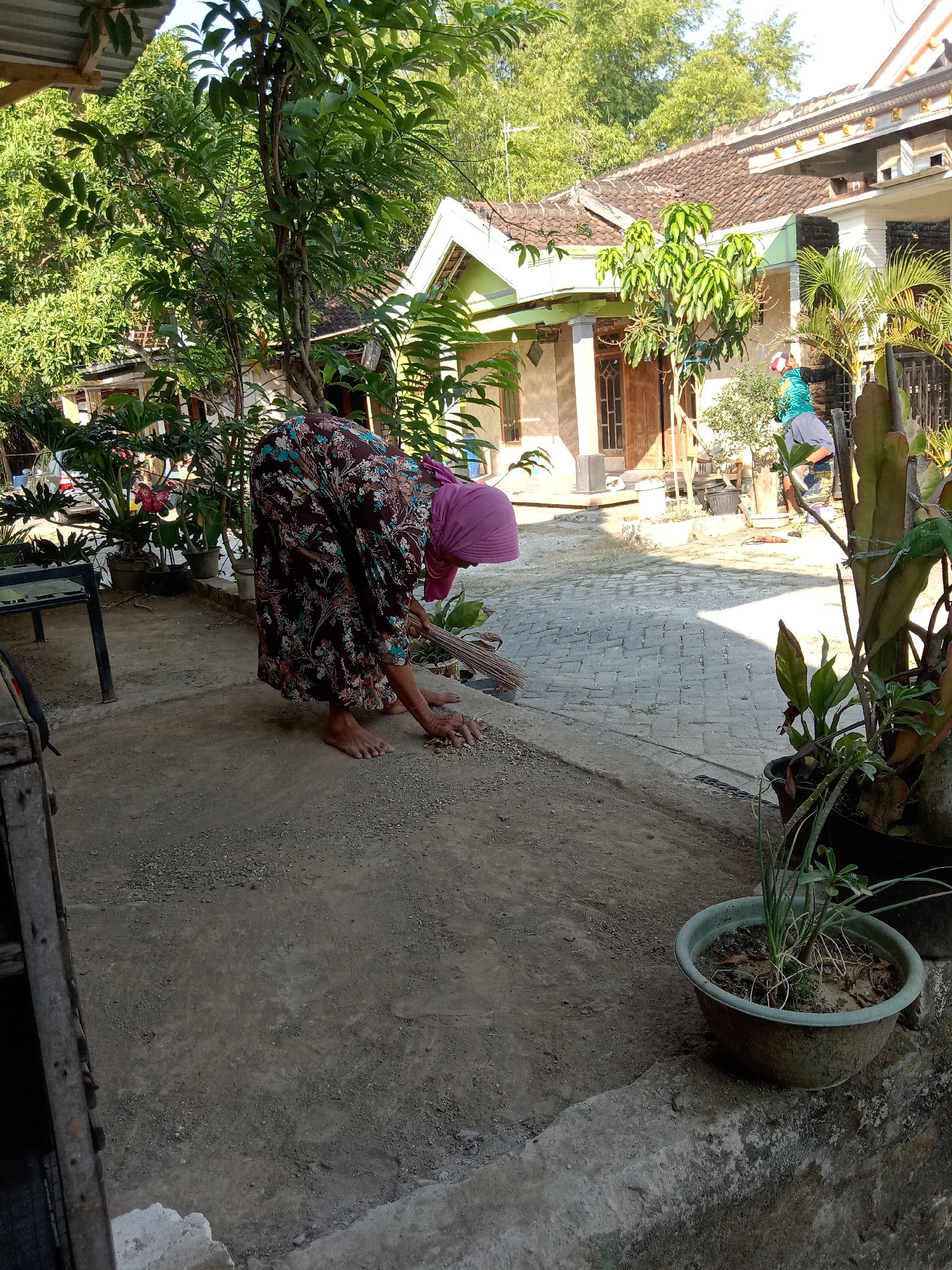 A woman wearing a pink headscarf and a floral-patterned dress is sweeping a gravel path outside a house in Stiaji, Indonesia. She is bent over, holding a broom and gathering debris. Several potted plants are visible around the property, along with a paved walkway and sections of building with tiled roofs. Another person, wearing a green shirt and dark shorts, is visible in the background, near the entrance of one of the houses. The scene appears to be a residential area bathed in sunlight.