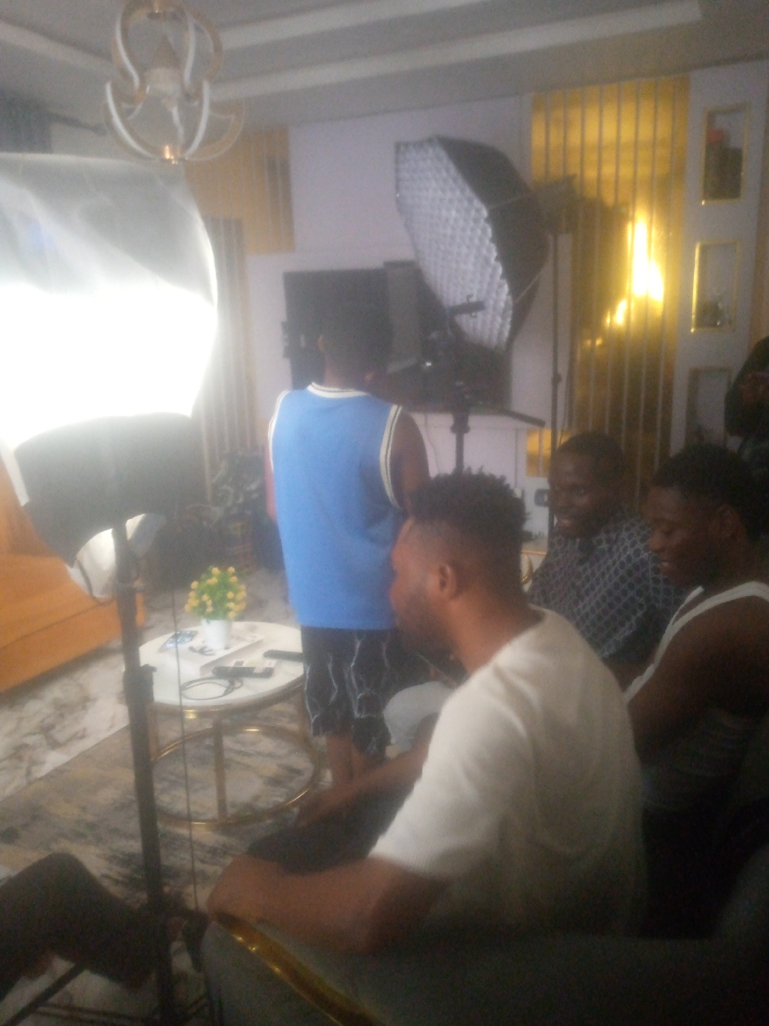 The image appears to be taken indoors, possibly in a studio or living room setting, within the city of Ikorodu, Nigeria. Several people are present: one person is wearing a blue jersey with a white trim, while two others are seen seated. Another individual has their back to the camera, and a man seated is wearing a patterned shirt, next to another person in a white tank top. A person behind him is cut off in the frame. Two large softbox lighting units are positioned around the scene, indicating a professional photoshoot or video recording. A small white round table with a flower arrangement and various items is present. The background features a television, a decorative wall, and an overhead light fixture. The floor is covered by a patterned rug.