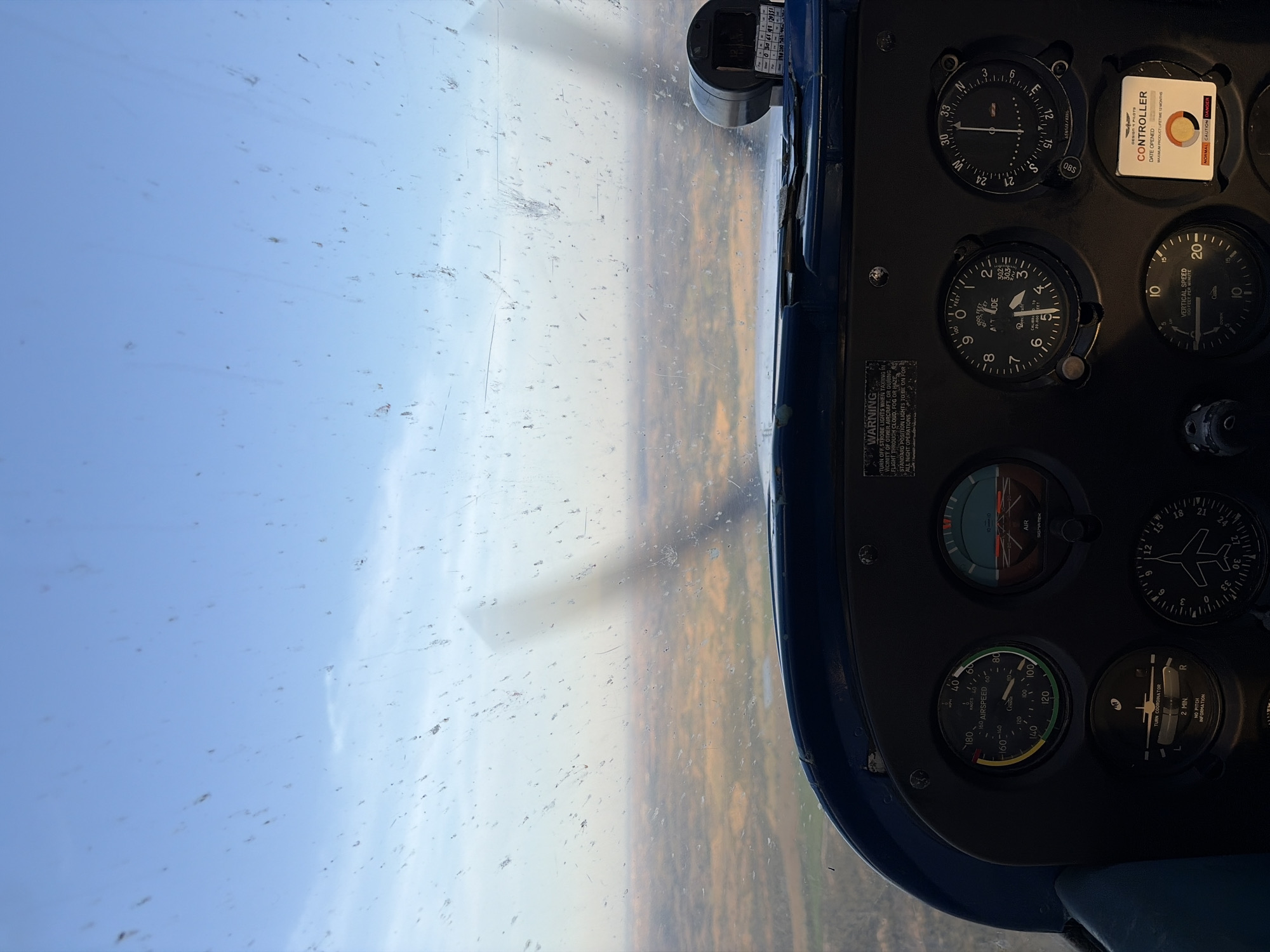 The image depicts an interior view of an aircraft cockpit during flight. The window on the left shows a view of the ground, likely a landscape with autumn-colored foliage and a partially clouded blue sky. The dashboard on the right contains various flight instruments, including an airspeed indicator reading approximately 80 knots, an altimeter indicating an altitude around 3,000 feet, a vertical speed indicator, a turn coordinator, and a navigation indicator. A "CONTROLLER" label is visible with a safety warning of "NORMAL CAUTION DANGER". Based on the landscape viewable through the window and the flight instrumentation, the aircraft is airborne, and the presence of an altitude indicator confirms that the aircraft is flying high above ground. Since the location is Villamantilla, Spain, it may be assumed the scenery includes the ground vegetation around that area. The details of individuals are absent in the image.