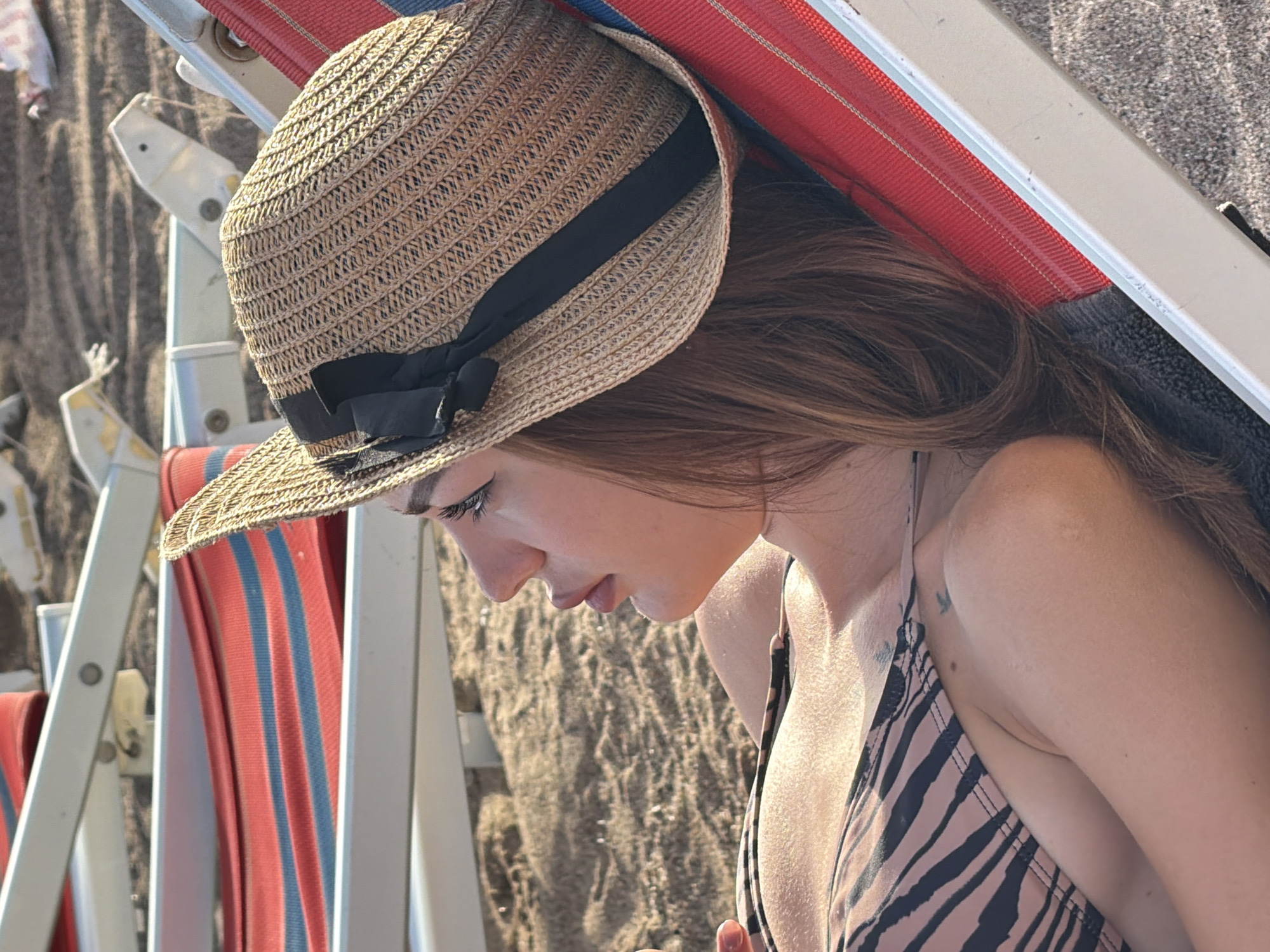 An overhead, slightly angled shot captures a woman relaxing on a beach chair in Boreti, Montenegro. The woman, positioned in profile facing left, wears a light brown straw hat adorned with a black ribbon and bow on the right side of her head. Her long, light brown hair falls over her shoulders. She is dressed in a swimsuit with a dark striped pattern, resembling zebra or tiger print, visible on her upper torso. Her eyes are closed or looking upwards. She is lying on a beach chair featuring a fabric sling with alternating red and blue horizontal stripes, supported by a light-colored metal or plastic frame. The sandy ground is visible beneath and around the chair. In the blurred background, additional beach chairs with light-colored frames are discernable.