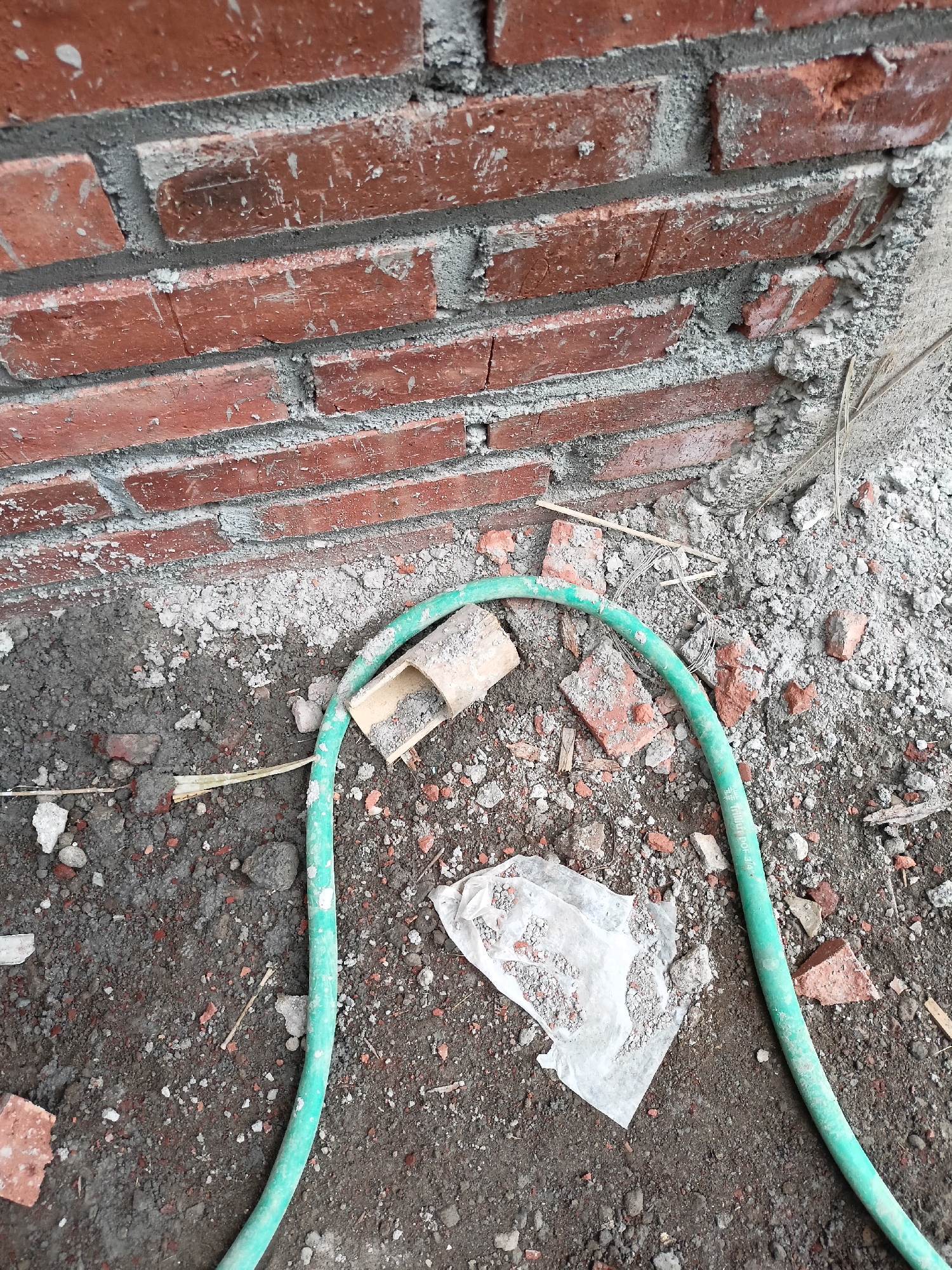 The image depicts a close-up, outdoor, ground-level view of a construction site at the Payaman location in Indonesia. A partially constructed brick wall, consisting of red bricks and grey mortar, forms the background. The ground is covered with dirt, rubble, and debris, including broken pieces of brick and scattered stones. A green garden hose is coiled on the ground, partially obscuring a crumpled piece of white plastic. Nearby, a light-colored, cylindrical object, possibly a piece of pipe or conduit, lies on the ground. There are no people or animals visible in the image. The lighting suggests it is daytime.