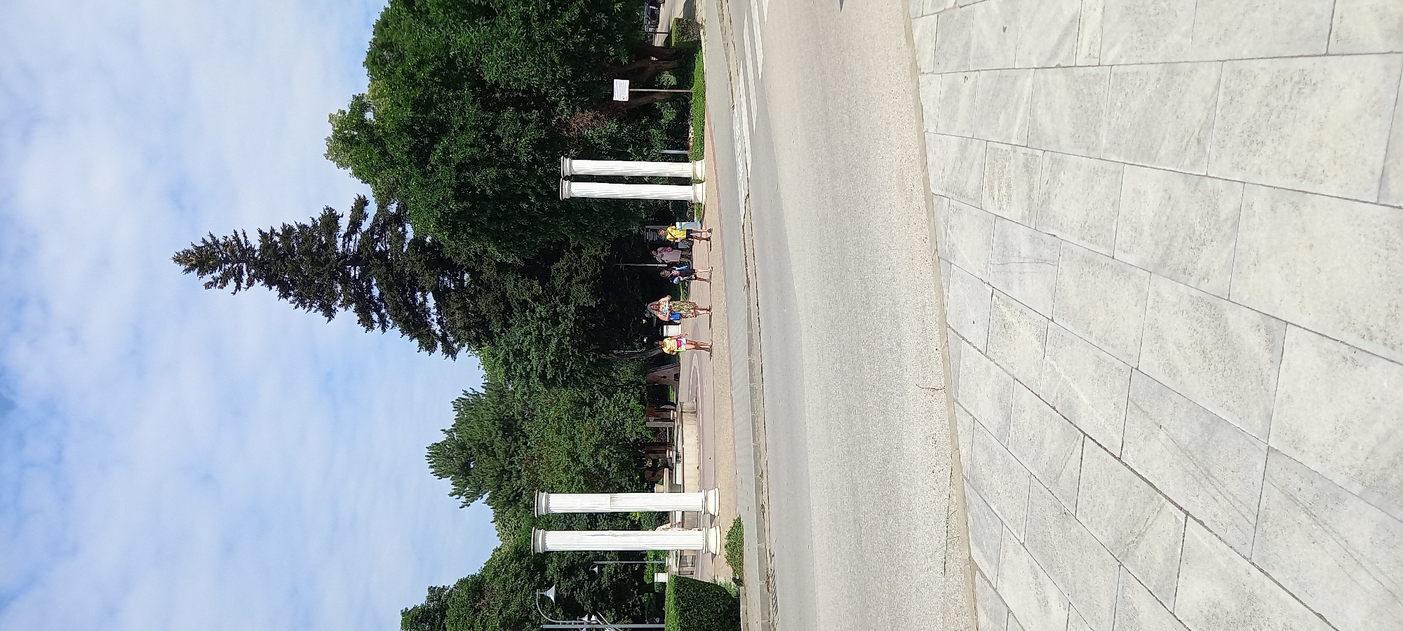 A paved area with a crosswalk and a street is in the foreground. Behind the street, there is a public space with several people, trees, and two large white columns. The columns are classical in design, with a fluted shaft and a capital. The people are walking and standing around, some of them appearing to be children. The trees are tall and lush, providing shade. In the background, there is a large evergreen tree, possibly a pine or fir, that stands out from the surrounding foliage. The sky is blue with some scattered clouds. The scene suggests a park or a public square in an urban environment. Specifically, this image captures the area near the "Drustar" hotel in Silistra, Bulgaria, showcasing a portion of the city's public spaces with notable architectural elements and natural surroundings.
