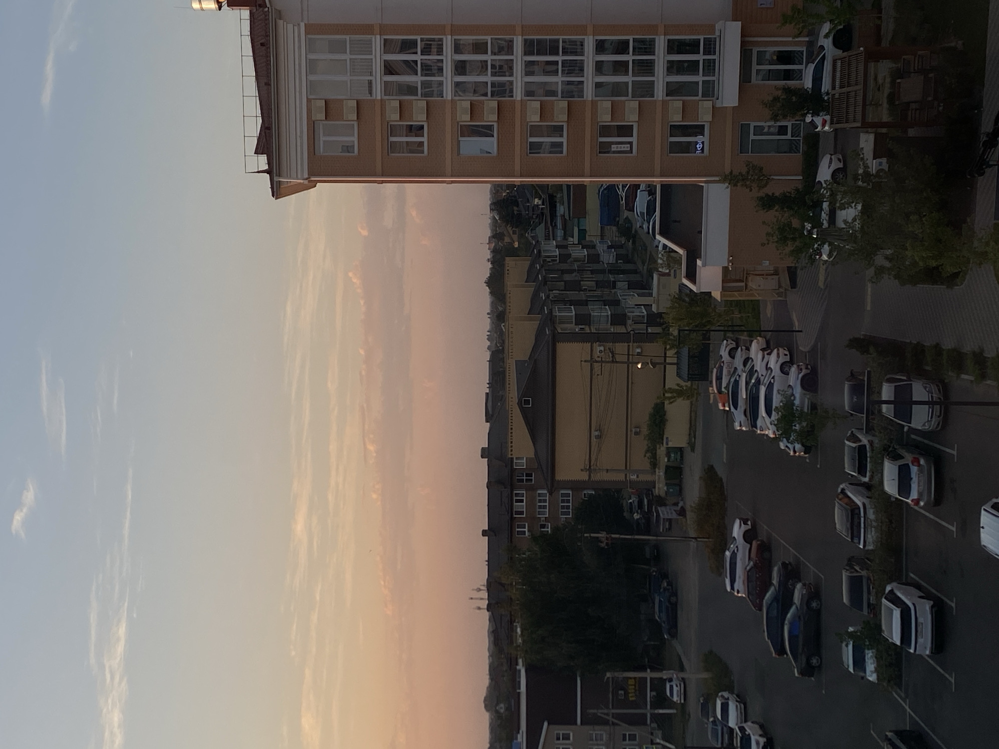 The image depicts an exterior view of a residential area in Zelenopol'skii, Russia, during twilight. The scene is dominated by a multi-story apartment building constructed with orange brick, featuring numerous windows and balconies. In the foreground, a parking lot contains a variety of cars, including sedans, SUVs, and smaller vehicles, parked in designated spaces. The ground surface of the parking lot appears to be paved. Adjacent to the parking lot are areas with trees and shrubbery, interspersed with some pedestrian walkways or paved paths. Other residential buildings of varying heights and architectural styles are visible in the middle and background, suggesting a moderately dense urban or suburban environment. The sky above is a gradient of light blue and soft pinkish hues, indicating the setting or rising sun. A few thin clouds are scattered across the sky. The overall atmosphere is calm and still, with no visible human activity.