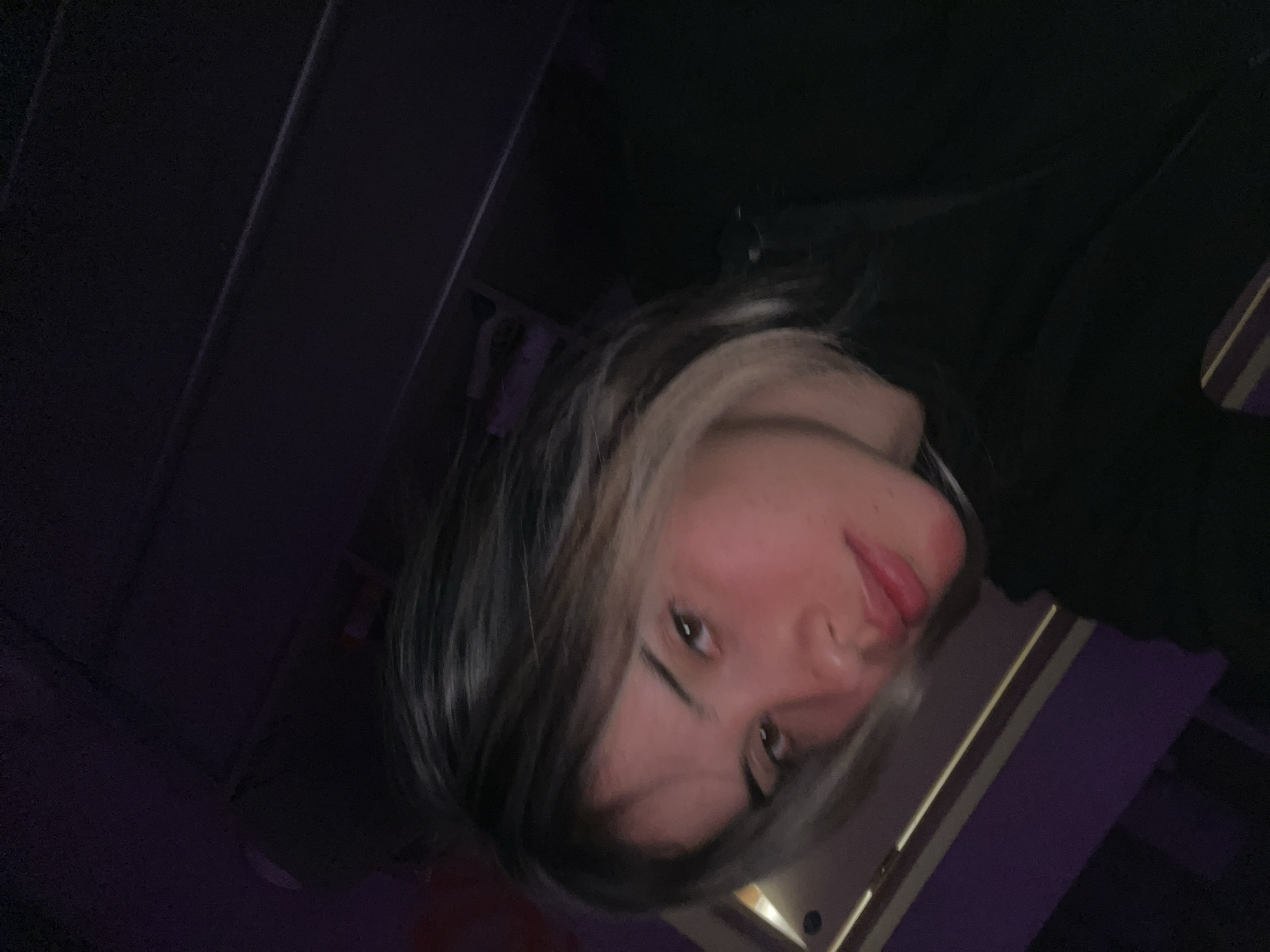 The image shows a close-up, vertically oriented, selfie-style portrait of a young person. The subject has dark hair with streaks of lighter color, particularly noticeable around the face. Their eyes are dark and looking towards the camera. The person's mouth is slightly open, with their tongue visible. The lighting is dim, creating shadows and highlighting the subject's features. The background is dark and appears to be an indoor setting with some vertical lines suggesting shelving or furniture. The color palette is dominated by dark tones with some purple hues visible, possibly from ambient lighting. There is no discernible text in the image. The overall mood is intimate and casual.