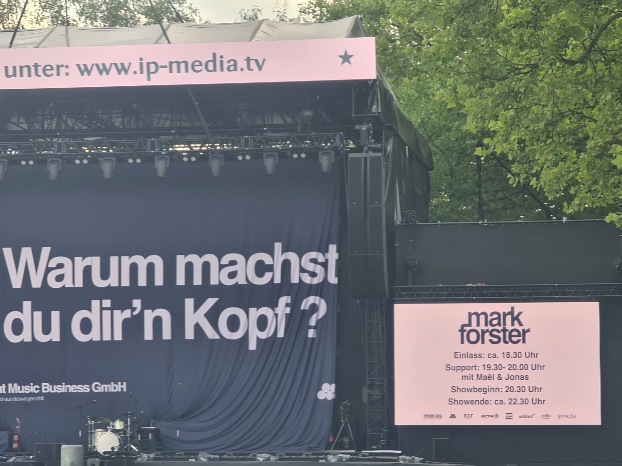 The image shows an outdoor stage setup, likely for a concert, with a large dark blue banner displaying white text in German that reads "Warum machst du dir'n Kopf?". Above this banner, a pink strip with white text and a star emblem indicates "unter: www.ip-media.tv". To the right of the stage, a large pink digital screen displays event information for "mark forster", listing entry time, support act details, and show start and end times. A drum set and other musical equipment are visible on the stage floor. Green trees and foliage are present in the background, suggesting an outdoor venue. The overall scene appears to be a public event in Klagenfurt, Austria.