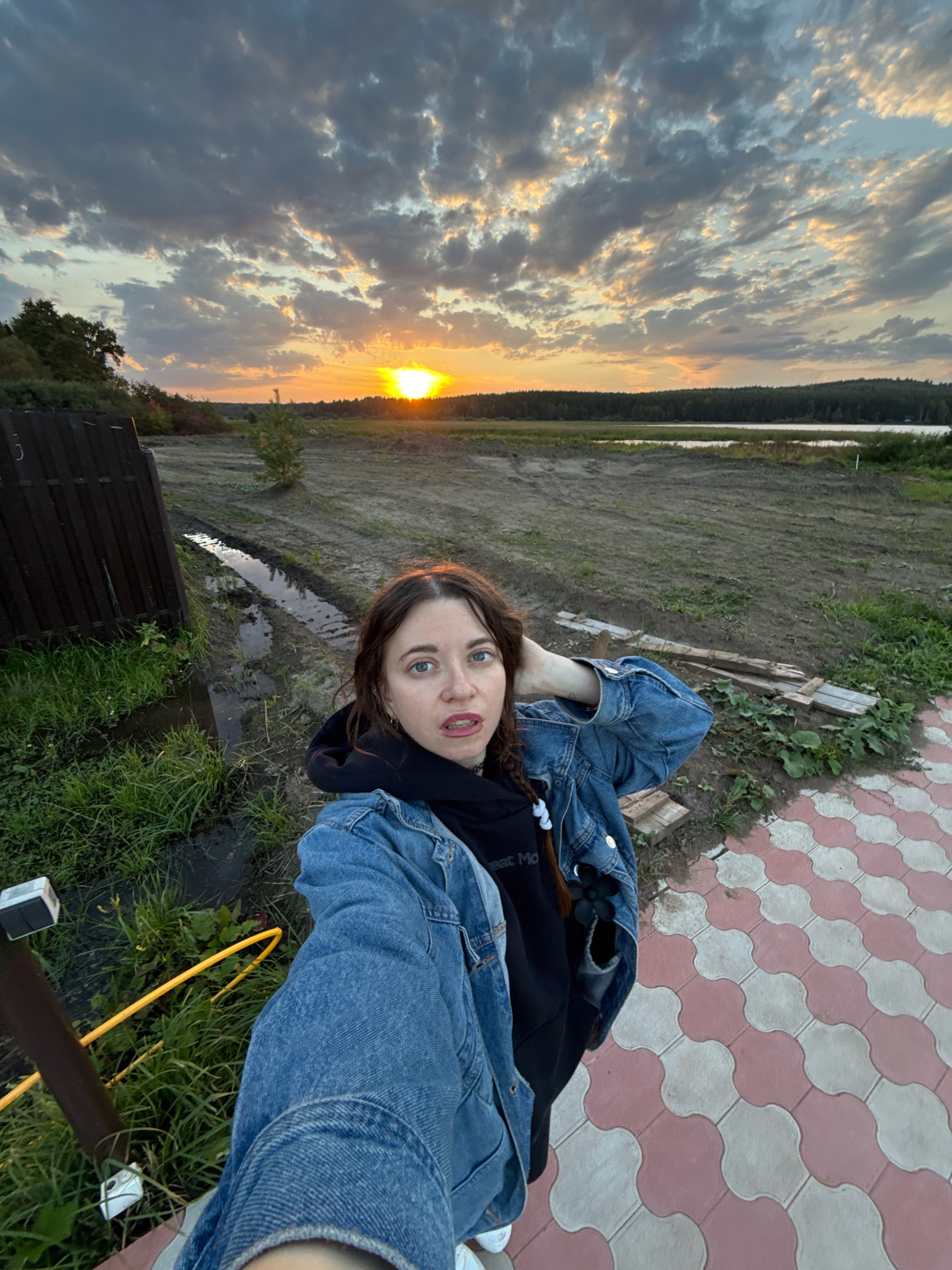This media file is a selfie of a young woman taken outdoors at dusk. She is wearing a denim jacket over a black hoodie, and her hair is in braids. She is looking at the camera with a surprised expression, her hand resting on the back of her head. The background shows a rural landscape with a wooden fence, a muddy patch with standing water, and a field leading to a line of trees and a body of water. The sky is filled with dramatic clouds illuminated by the setting sun, casting an orange glow. The woman is standing on a decorative paved area in the foreground. The visible text on her hoodie reads "eat Mo". The setting appears to be the countryside in Pakli, Russia. The overall mood is casual and contemplative, with the natural beauty of the sunset contrasting with the somewhat unkempt foreground.