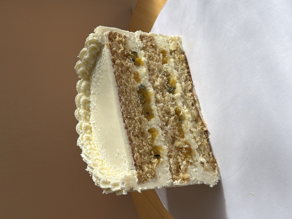 A single slice of multi-layered cake is centered on a white, subtly patterned surface. The cake consists of three visible layers of light brown sponge cake, alternating with three layers of white cream or frosting. Embedded within two of the white frosting layers are distinct sections of yellow-orange fruit pulp containing dark seeds, consistent with passion fruit filling. The top surface of the cake slice features a smooth white glaze, bordered by a piped decorative edge of white frosting, possibly adorned with shredded coconut flakes. The exterior side of the cake is coated in white frosting. Illumination suggests a single light source from the right, casting a soft shadow on the left side of the cake. The background is a uniformly plain, light brown or beige wall, out of focus. The image provides no discernible details to indicate its specific location within the city of Tel-Aviv, Israel, or any other geographical area. There are no individuals present, nor any actions or interactions depicted.