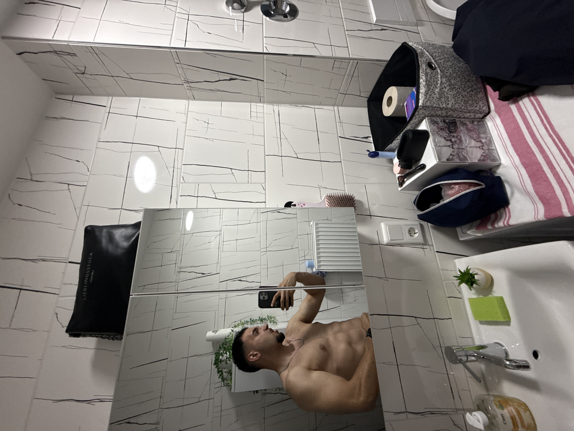 A young adult male, shirtless, is taking a mirror selfie in a bathroom. He is standing to the left of the frame, flexing his bicep and making a grimacing face. He is wearing a silver chain necklace and a watch on his left wrist. The bathroom features white tile with black line patterns. To the left, a sink with a chrome faucet and a green sponge are visible. Above the sink, a mirrored cabinet reflects the man. A black clutch with "LIEBLINGSSTÜCK" embossed on it sits on top of the cabinet. On the right side of the frame, various bathroom items are organized on a table, including a black storage bin with a roll of toilet paper and a small plant. The image was taken in a bathroom in Garmisch-Partenkirchen, Germany.