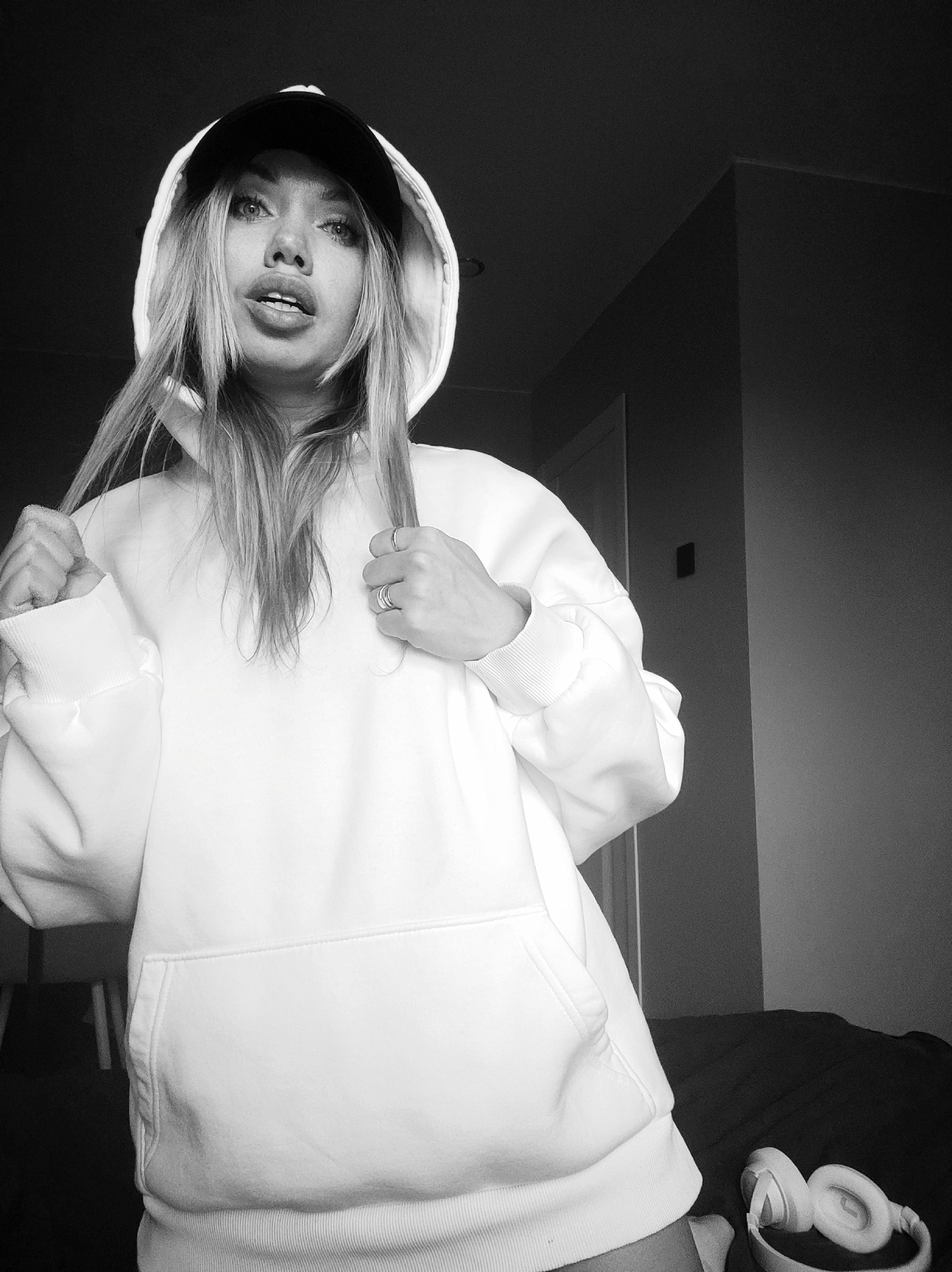 A black and white, low-angle photograph captures a young person indoors, looking directly towards the camera. The subject, likely female, is wearing an oversized, light-colored (appearing white or light grey) hoodie with the hood up, partially covering a dark baseball cap worn underneath. Her long, light-colored hair falls forward around her face.
Her expression features wide eyes and lips slightly parted. She holds strands of her hair with her left hand, while her right hand, adorned with multiple rings, rests on her chest, subtly pulling at the hoodie fabric. The hoodie has ribbed cuffs and hem, and a large kangaroo pocket on the front.
The background is simple and mostly dark, suggesting a dimly lit room. A dark wall corner and a ceiling are visible in the upper right. To the left, a hint of furniture and possibly a doorframe can be seen, along with a dark rectangular object mounted on a wall. In the lower right corner, a pair of white over-ear headphones rests on a dark surface, possibly a bed or sofa.
The image appears to be a posed shot, focusing on the individual and her attire within a minimalist setting. The photograph lacks any visible text. The location context for this scene is Votkinsk, Russia.